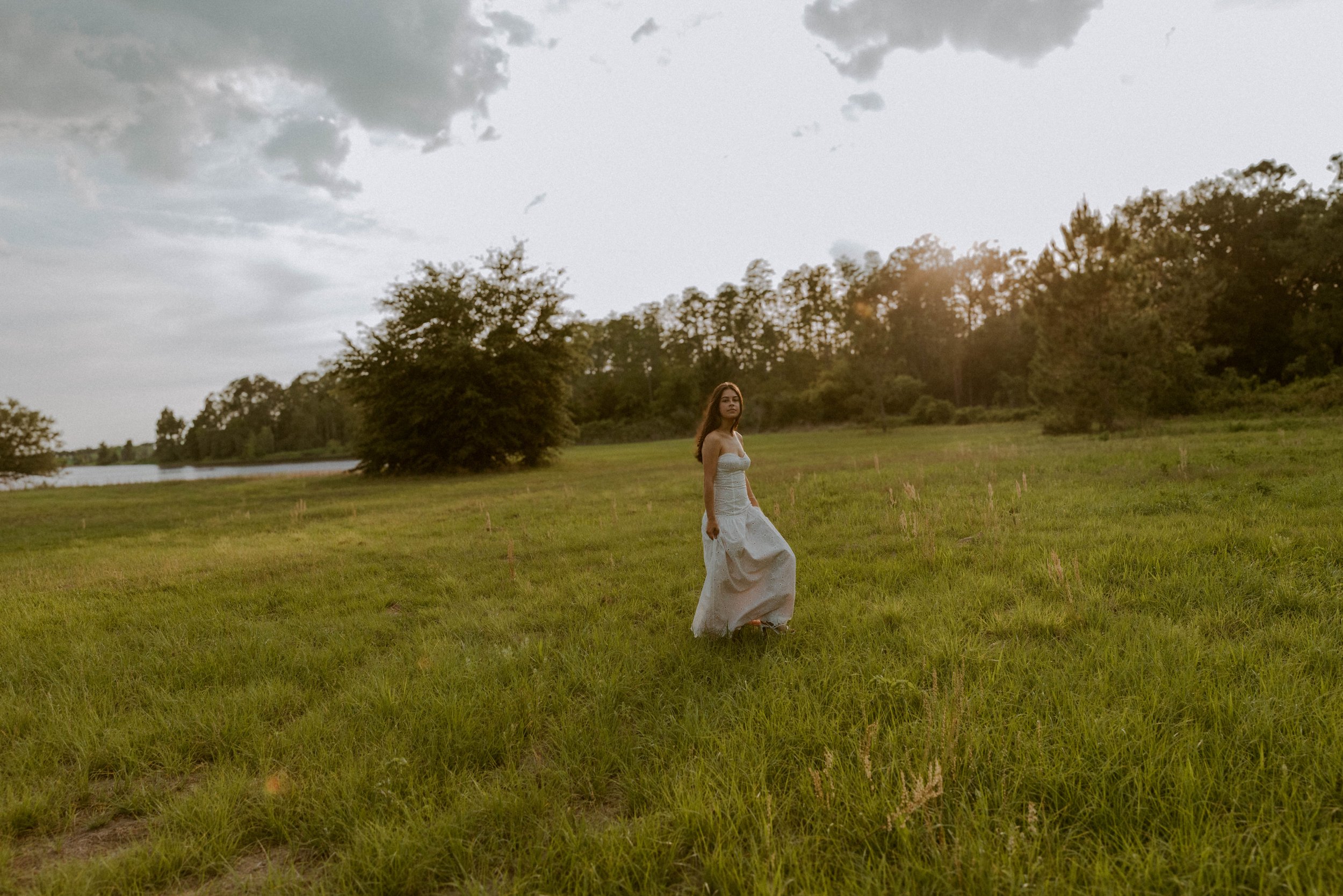 A woman in a long white dress walking across a grassy field with trees in the background, on a cloudy day.