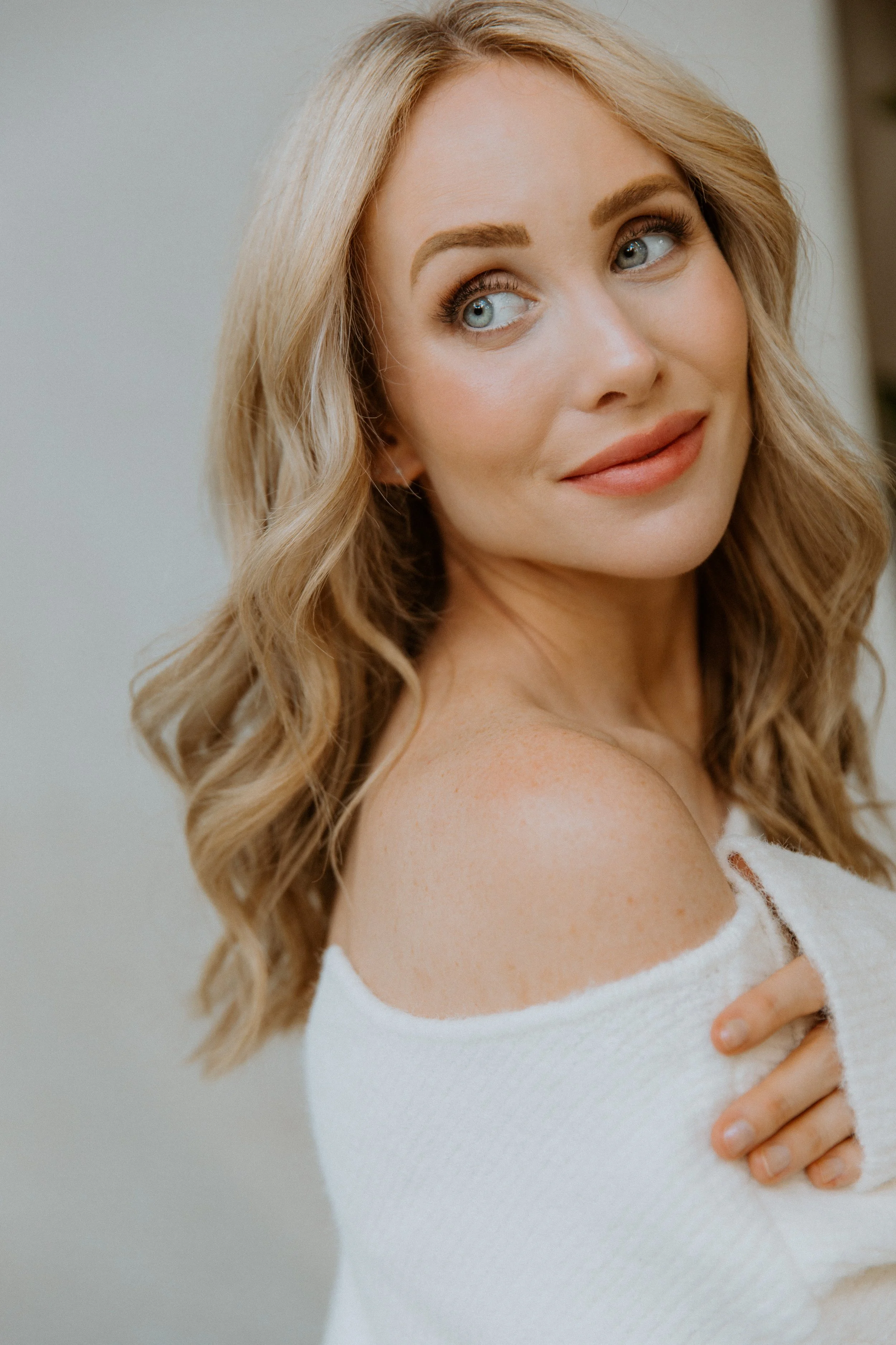 Close-up of a woman with blonde curly hair, blue eyes, and a light skin tone, wearing a white off-shoulder top, with a gentle smile, touching her shoulder with her hand.