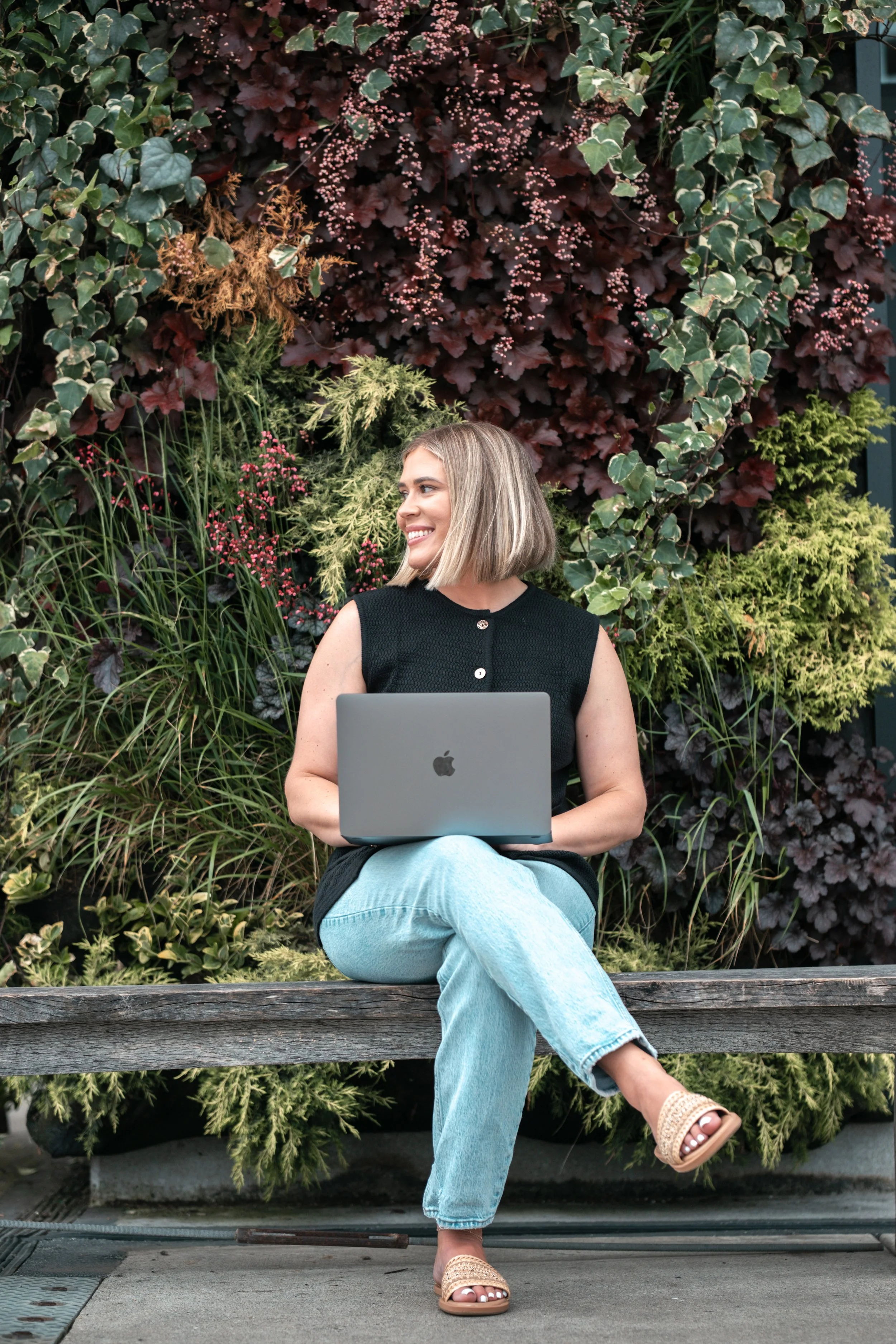 A young woman with shoulder-length blonde hair, wearing a sleeveless black top and light blue jeans, is sitting on a wooden bench outdoors, holding a silver MacBook in her lap and smiling while looking to her right. Behind her is a lush wall of green, burgundy, and reddish leaves and plants.