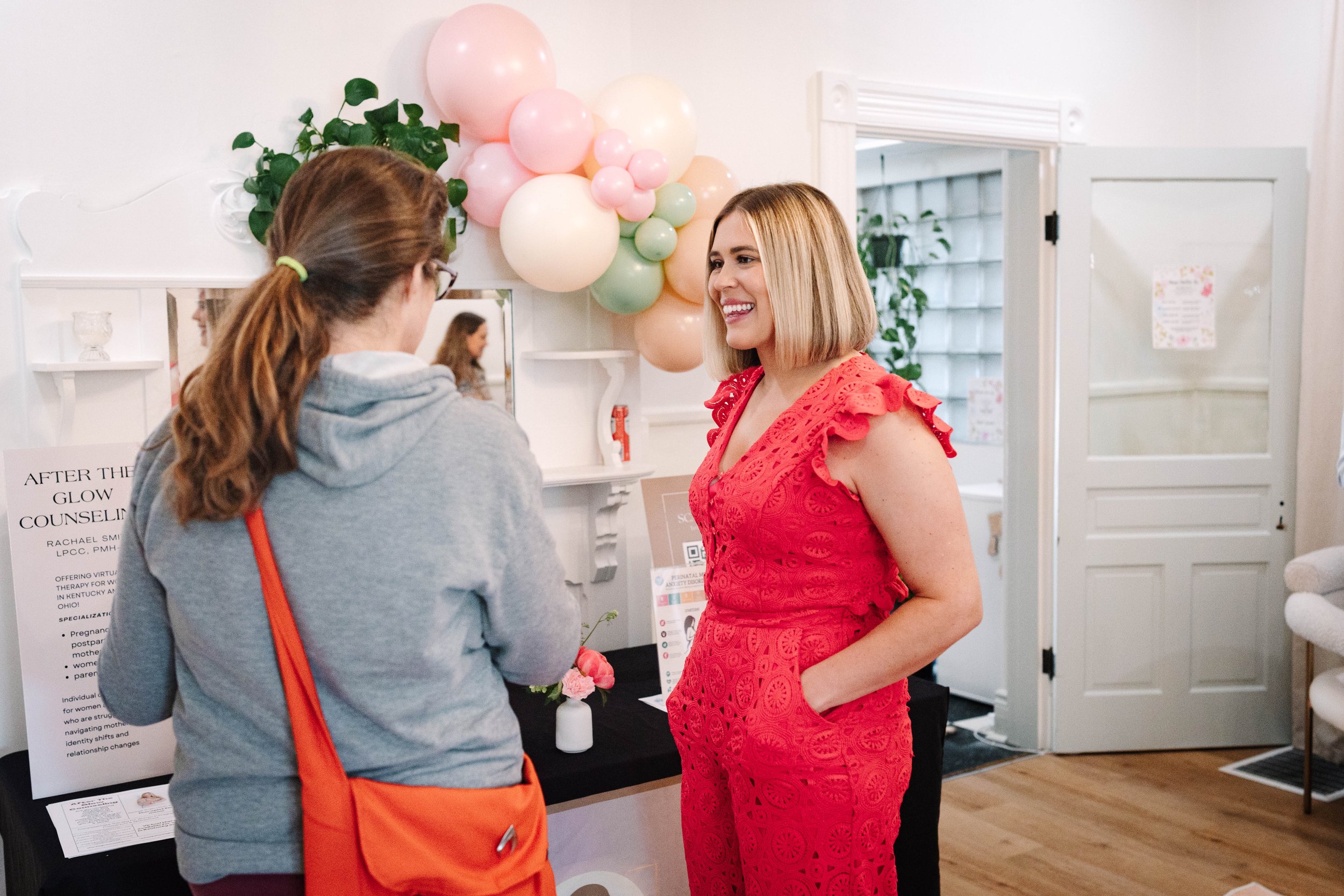 Woman in a red dress smiling and talking to another woman in a gray hoodie at an indoor event with balloons and display table.