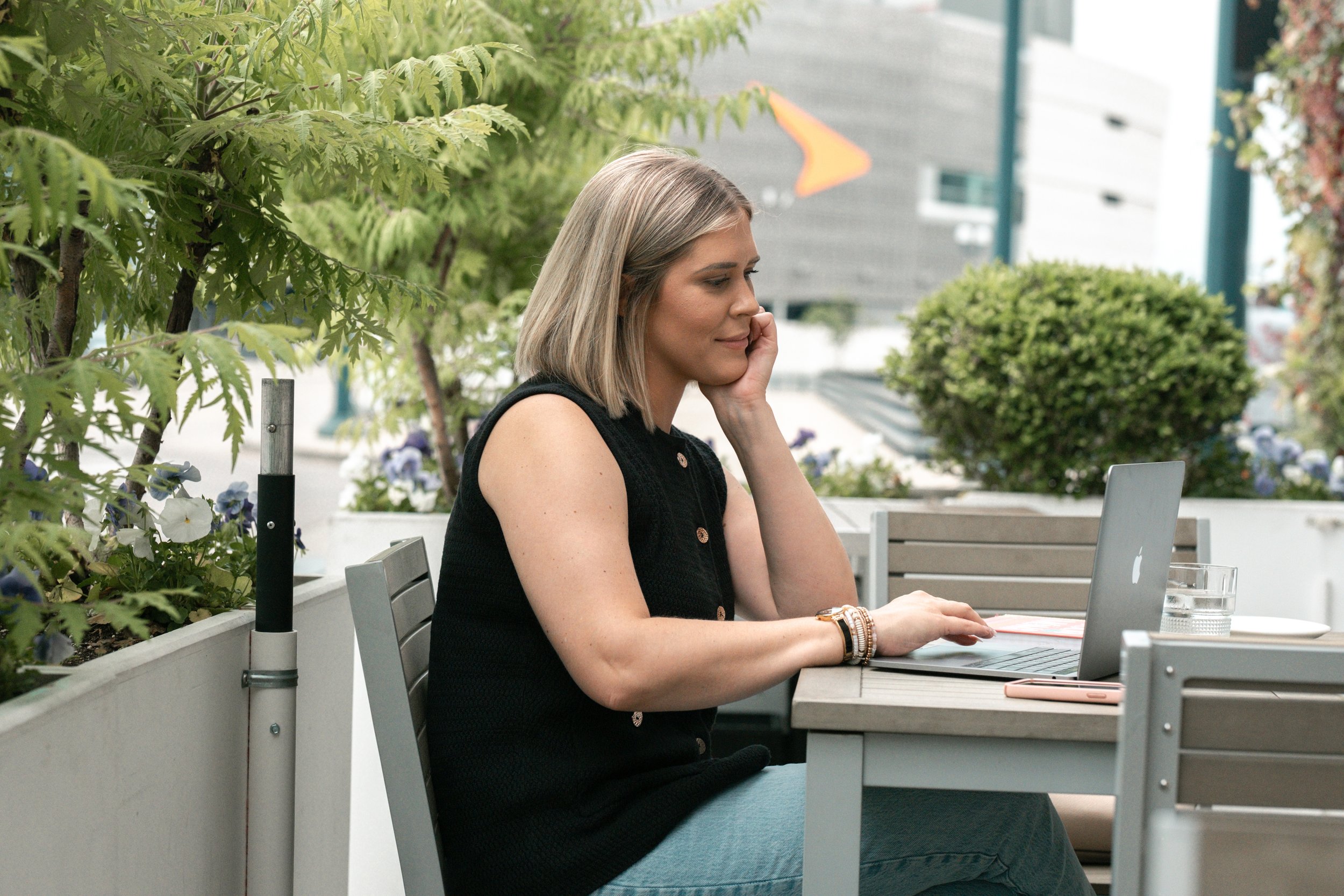 A woman sitting at an outdoor patio table, working on a silver MacBook laptop, with a glass of water and a smartphone nearby, surrounded by green plants.