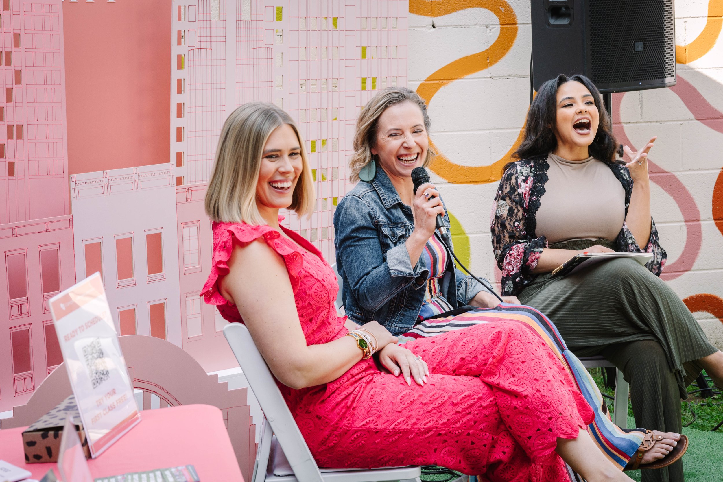 Three women sitting in chairs at a panel discussion or event, smiling and laughing with one woman holding a microphone. The backdrop has abstract colorful designs on a brick wall.