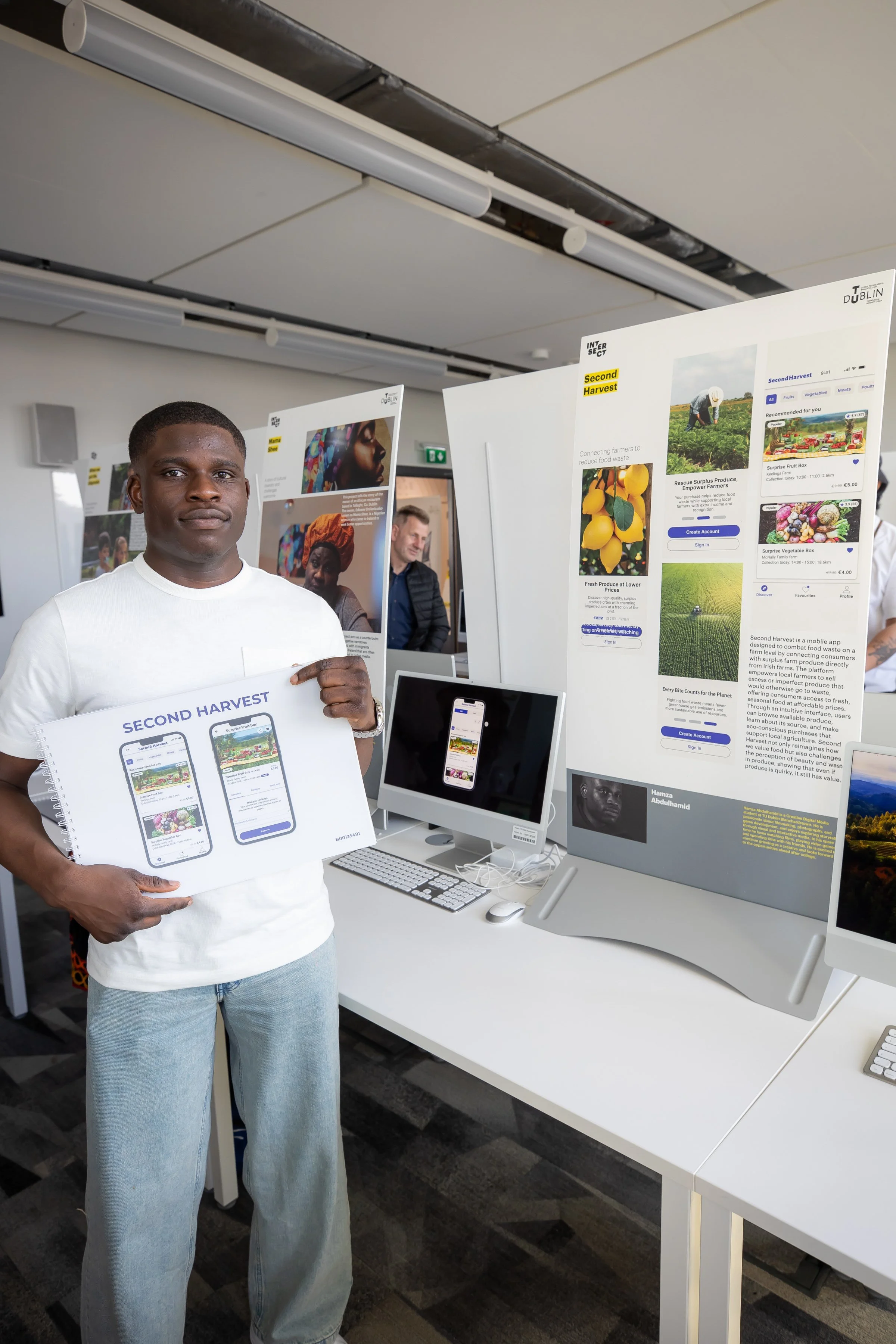 A man holding a presentation board for the Second Harvest project at an exhibition or conference, with display screens and posters about the project on the tables behind him.