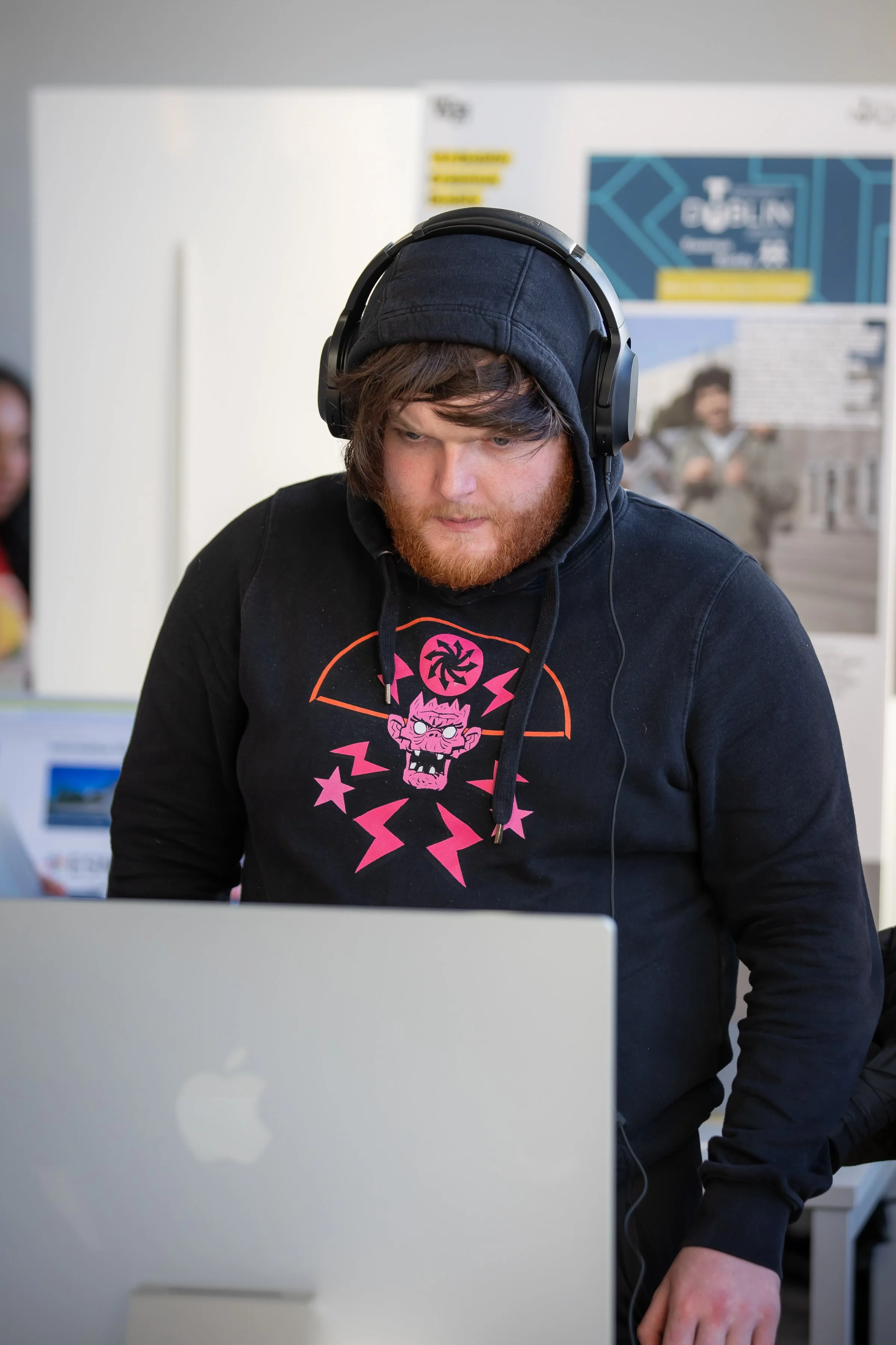 A young man with a red beard and brown hair wears headphones and a black hoodie with a pink graphic, working on a silver Apple MacBook computer in an indoor setting.