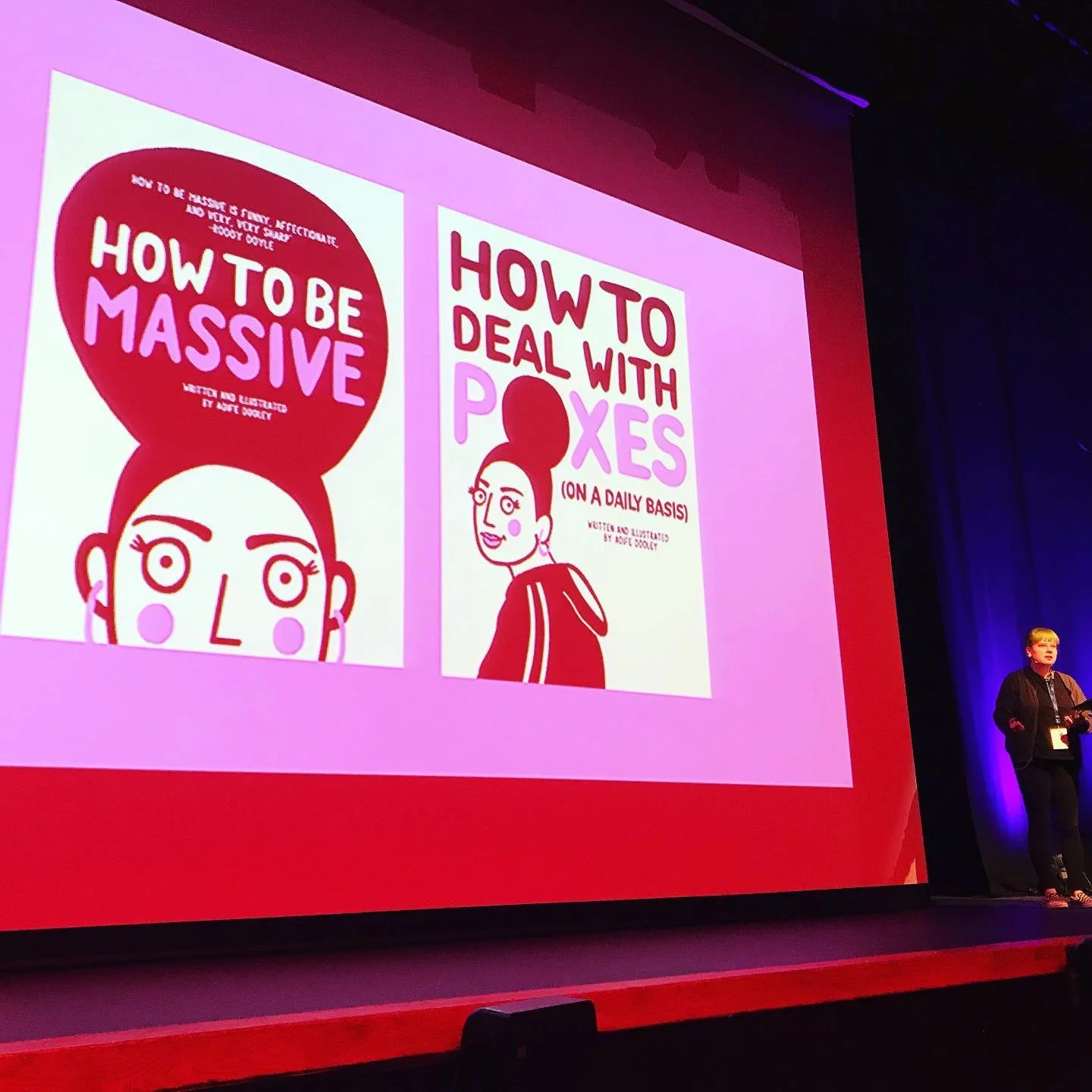 A woman presenting on stage in front of a large screen displaying two book covers, one titled 'How to Be Massive' and the other 'How to Deal with Poxes (on a Daily Basis)', both with illustrations of women.