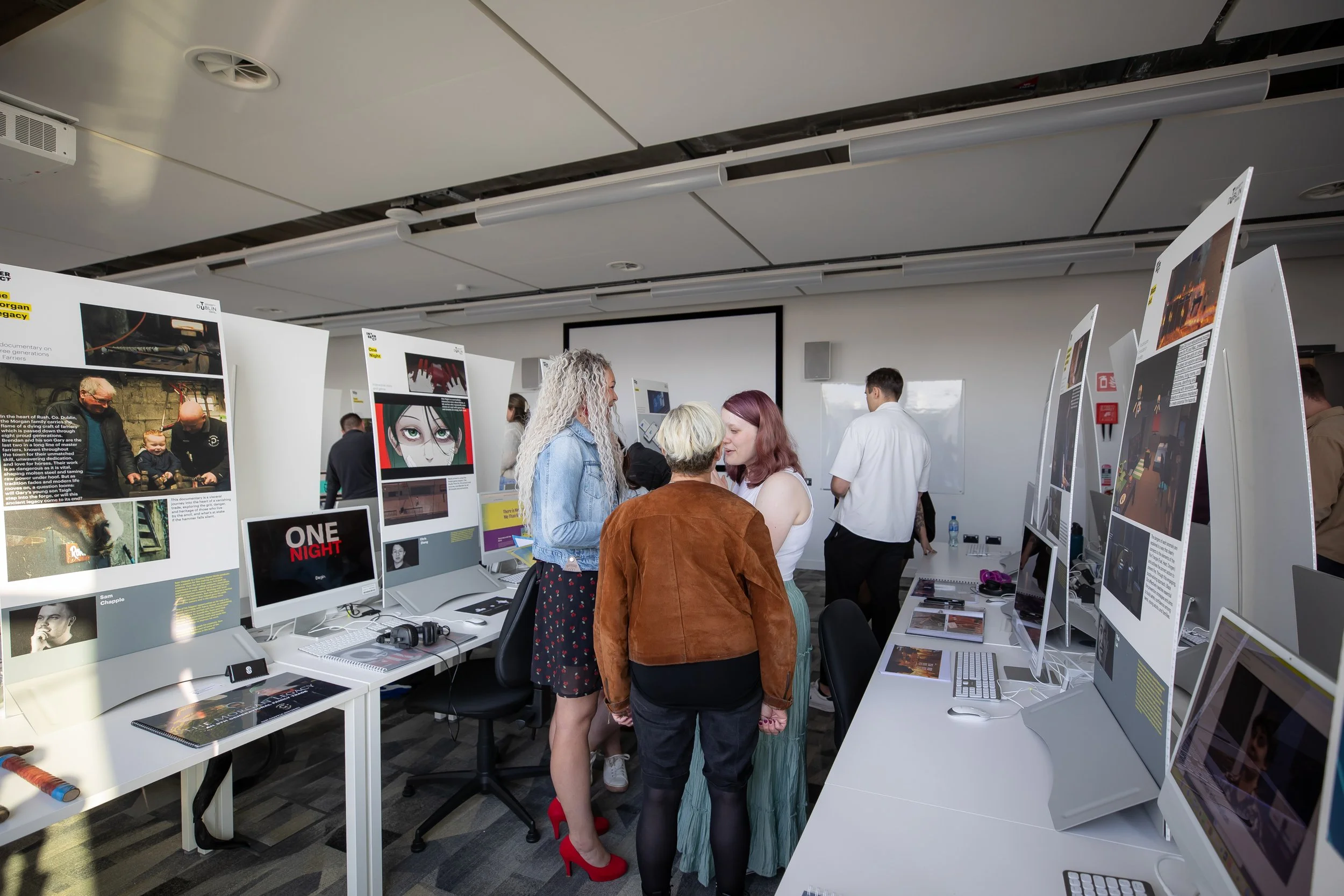 People viewing and discussing art and design displays in an exhibition room with computers and posters.
