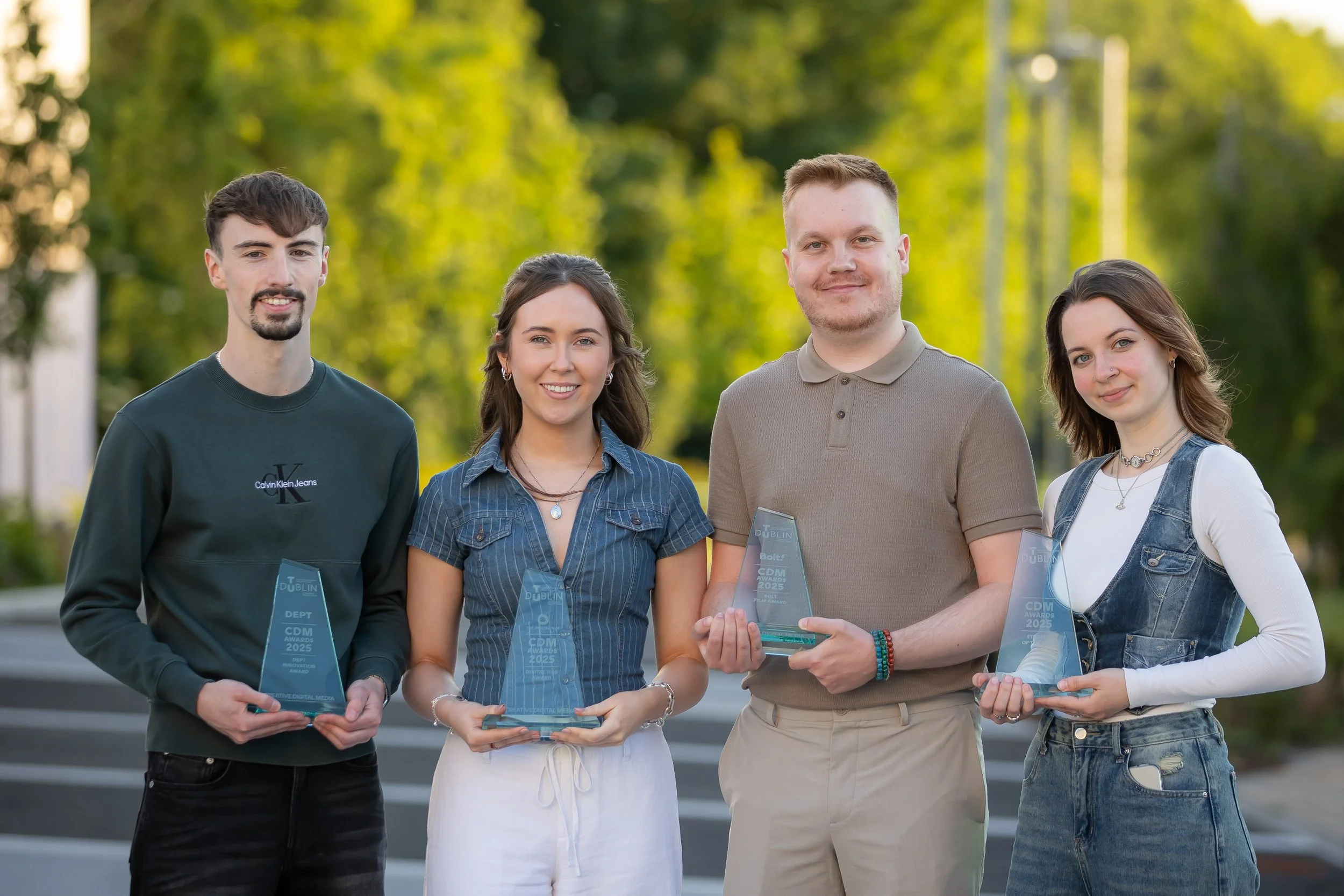 Four young adults standing outdoors in a park, holding award trophies and smiling, with green trees in the background.