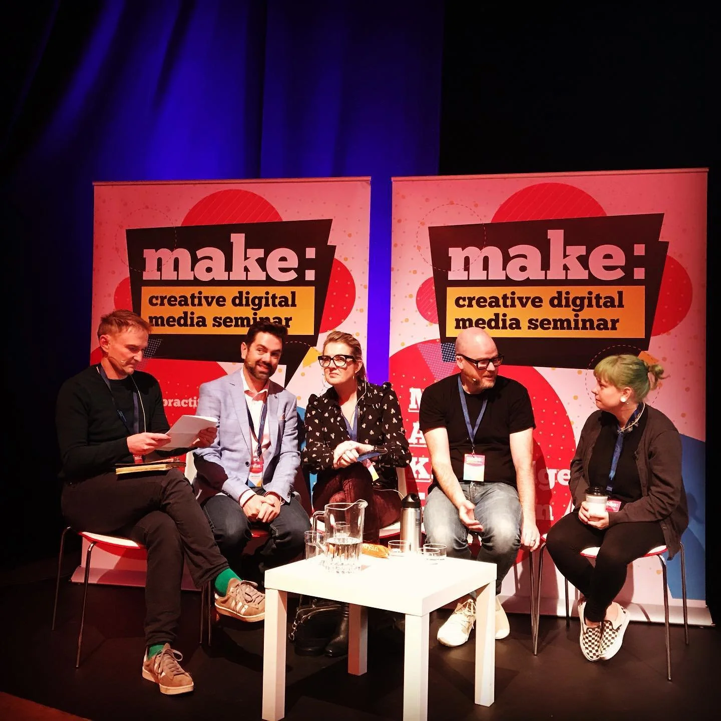 Five people sitting on stage at a digital media seminar, with two large banners behind them that read "make: creative digital media seminar" and a table with pitchers and cups in front of them.