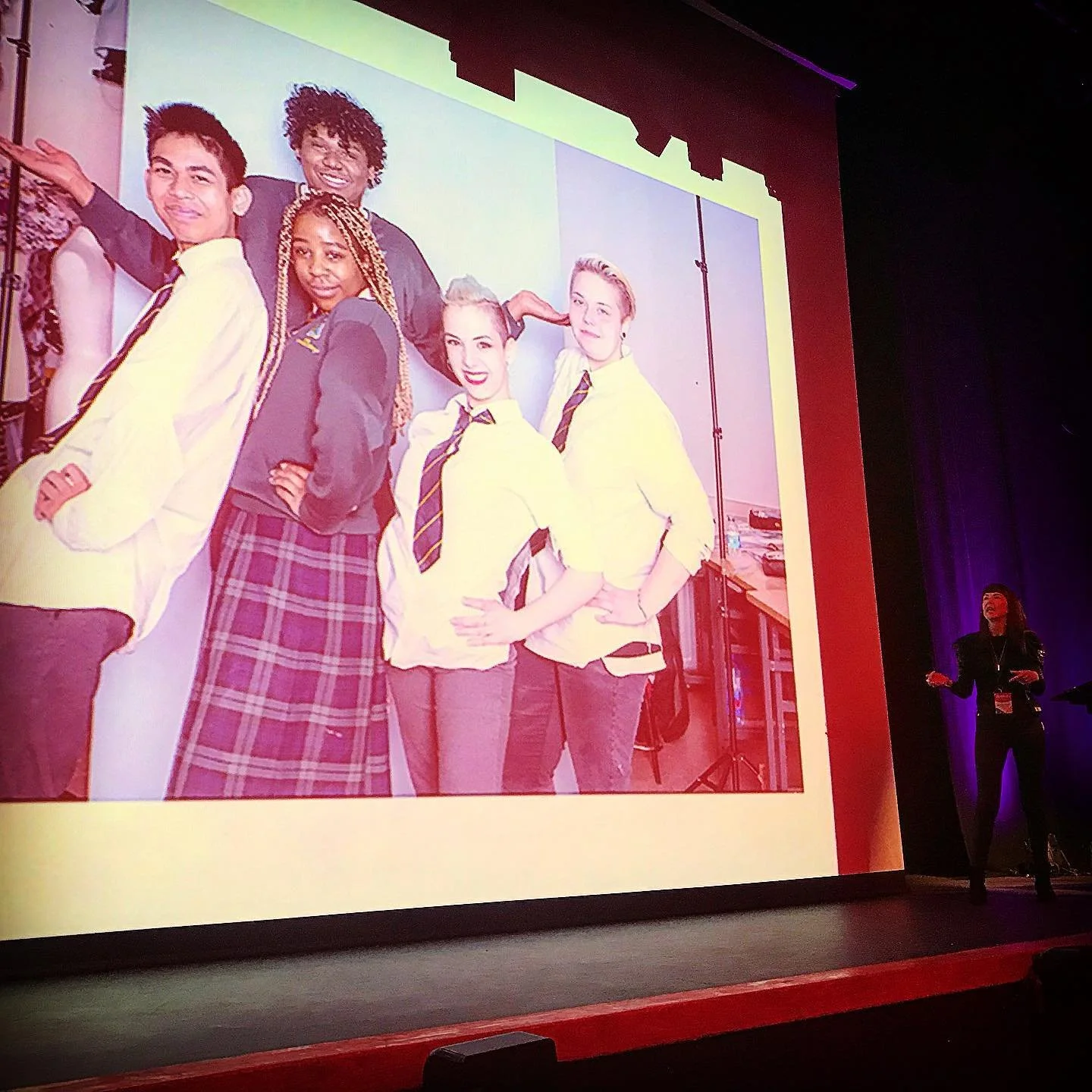 A large projection of a group of five young people, dressed in school uniforms, standing together and smiling. The background appears to be a school setting. A woman is standing on a stage to the right, giving a presentation.