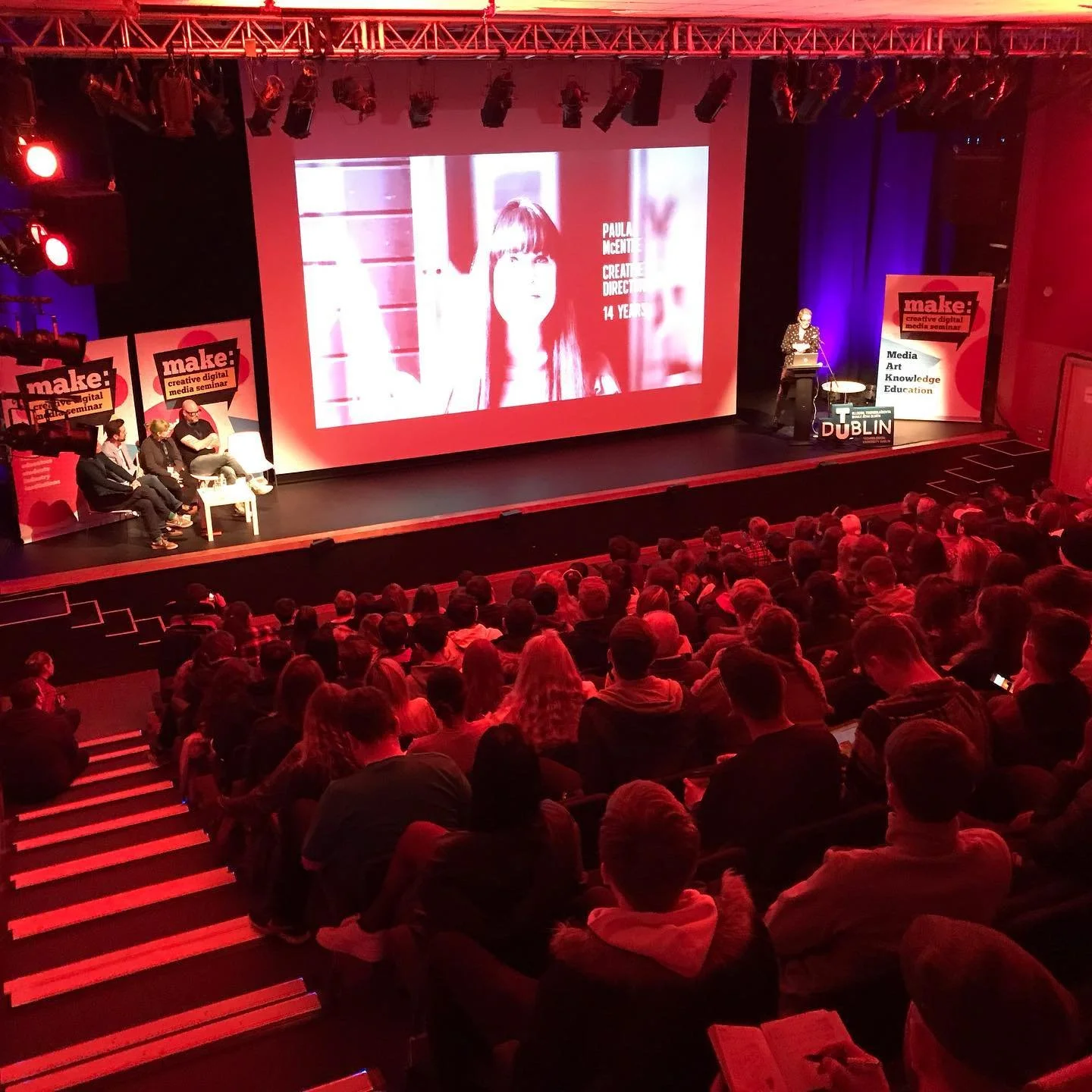 A large audience seated in a darkened theater watching a presentation on stage. The stage features a large screen displaying a woman's face and text, with four panelists sitting to the left and a speaker at a podium to the right. The event is titled
