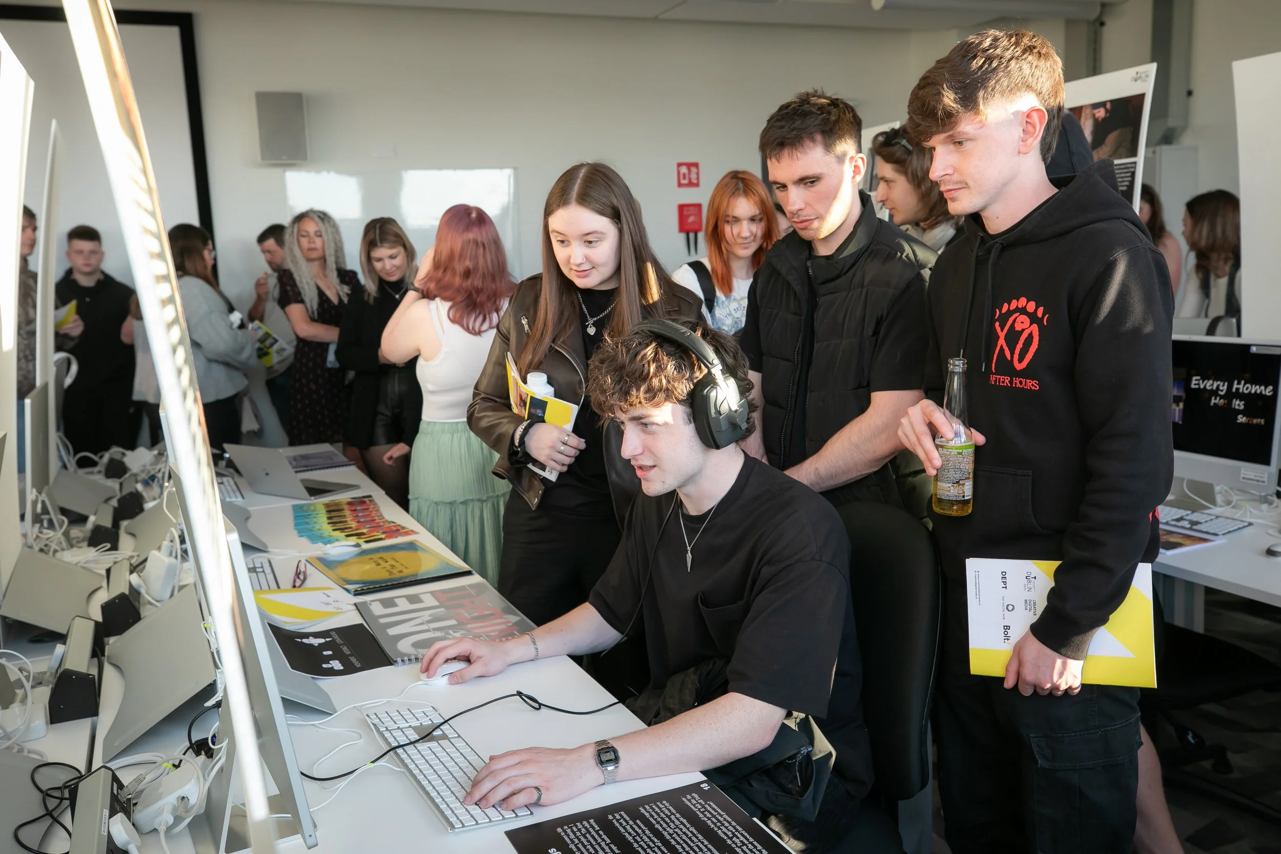 A group of people gathered around a young man wearing headphones and working on a computer at a busy technology event or exhibition.