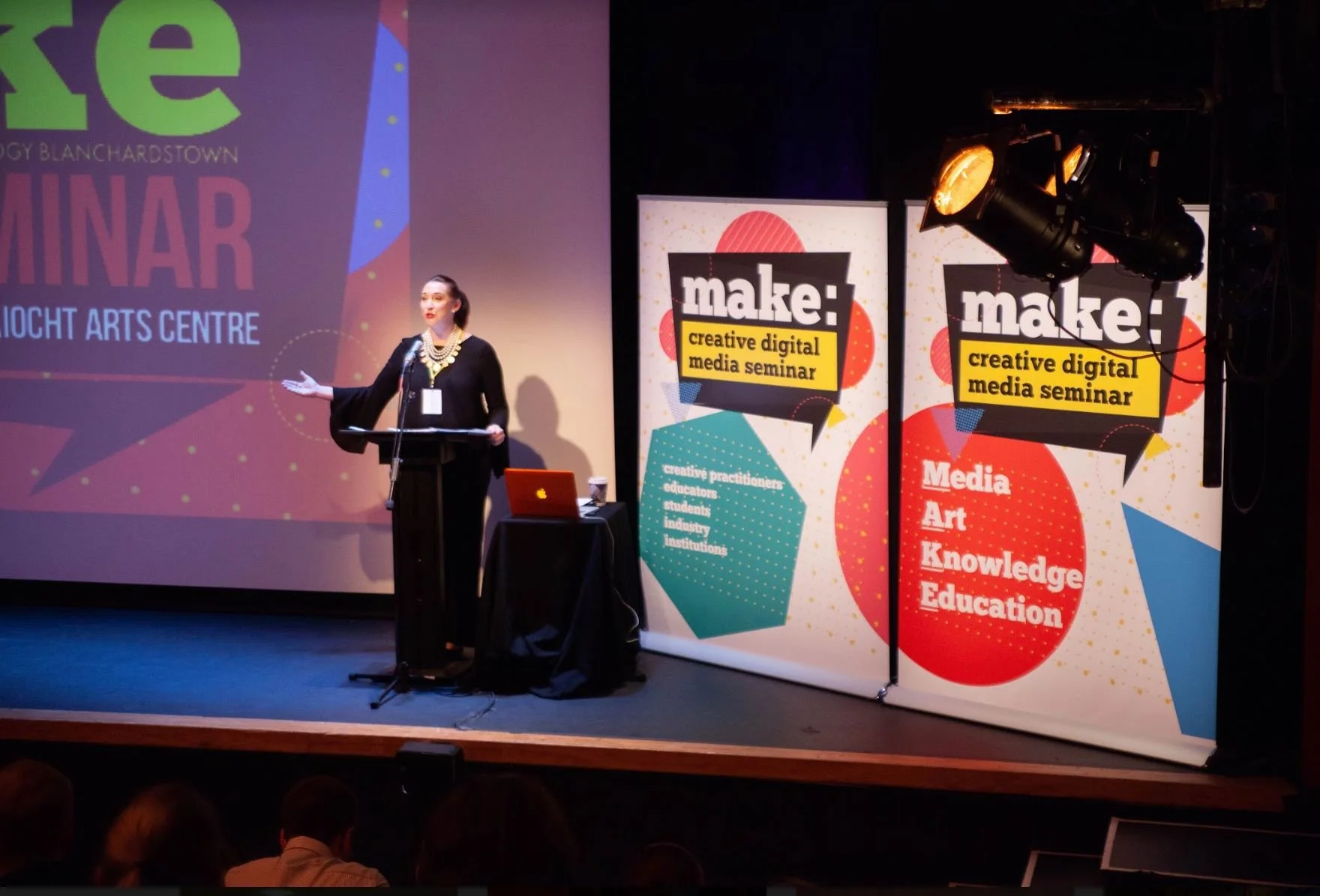 A woman wearing black, standing at a podium with a laptop, speaking at a seminar. Behind her are two vertical banners with colorful designs and the words 'make: creative digital media seminar.' A large projection screen displays similar seminar-relat