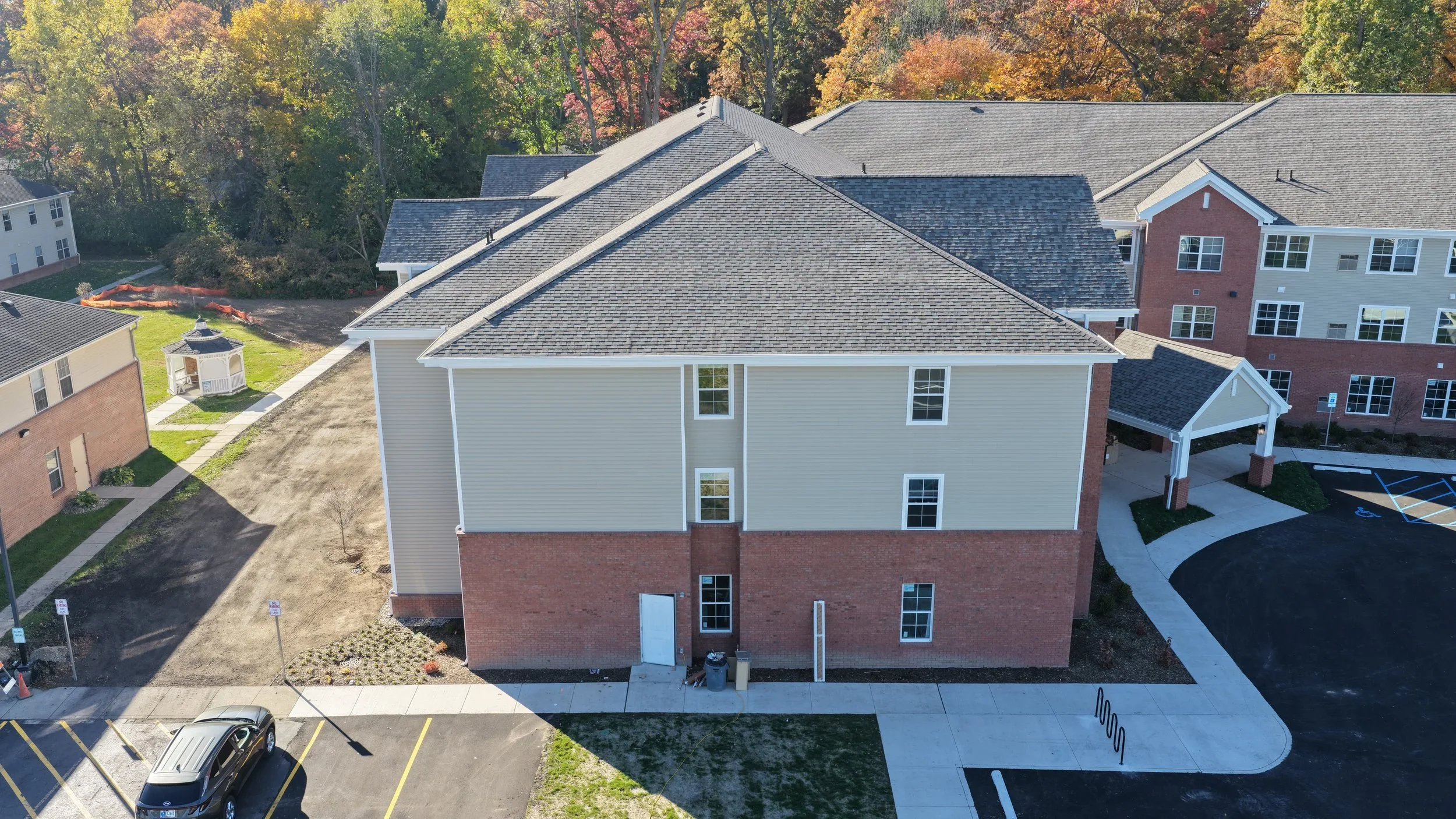 Aerial view of a multi-story residential apartment building with beige siding and brick lower walls, surrounded by parking lots, sidewalks, and other residential buildings, with trees in the background.