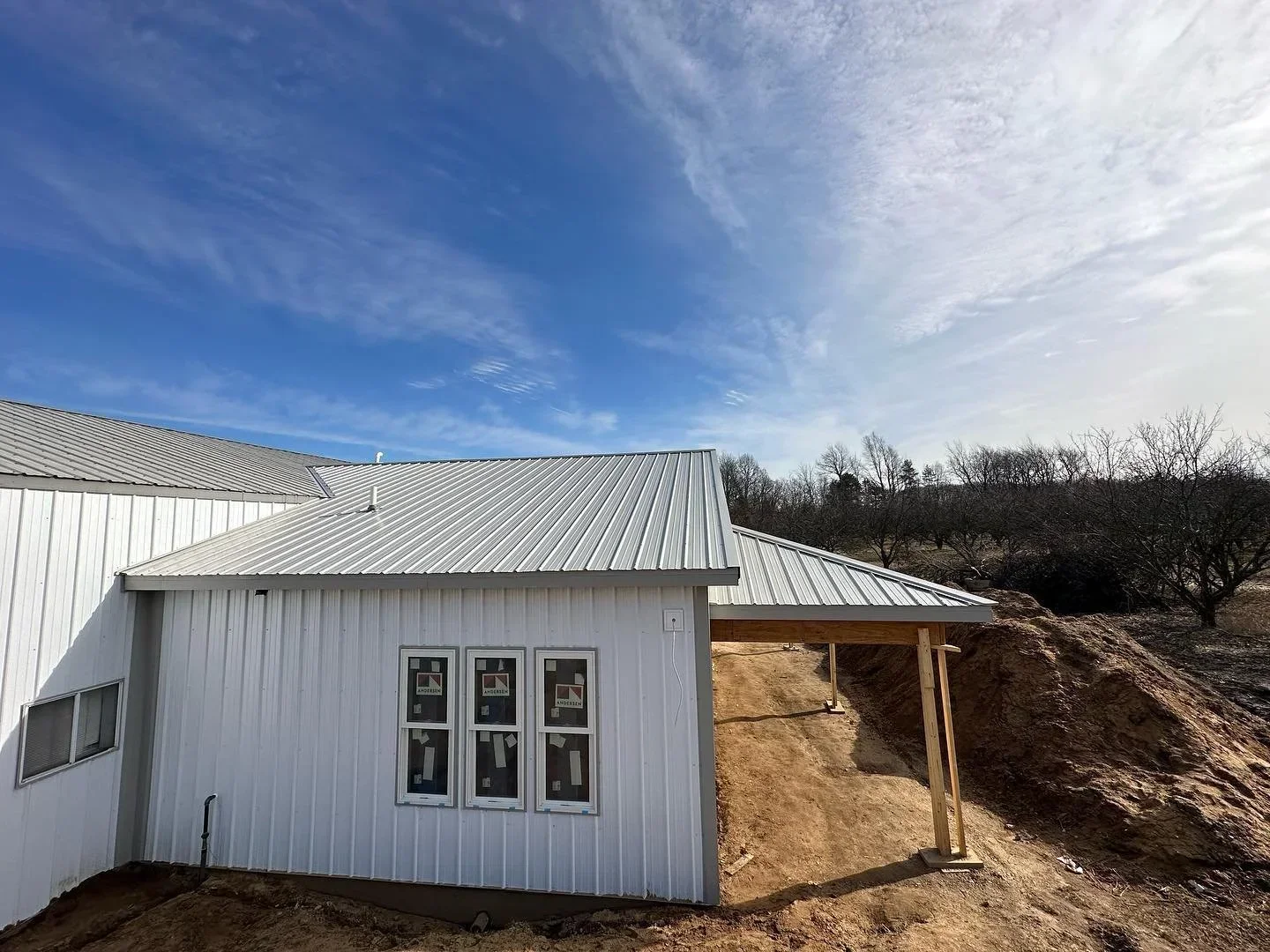 White metal building under construction with a sloped roof, three windows, and bare trees in the background on a clear day.
