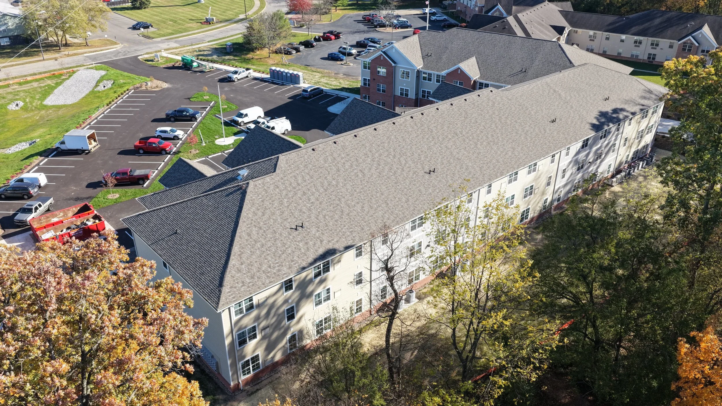 Aerial view of an apartment complex with parking lots, trees with autumn foliage, and green surroundings.