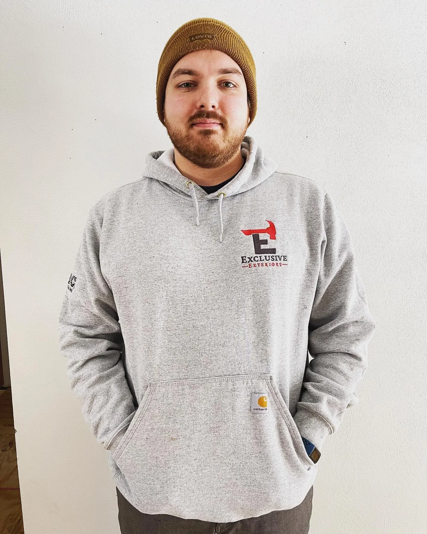 A man wearing a yellow beanie and a gray hoodie with a logo, standing against a plain light wall.