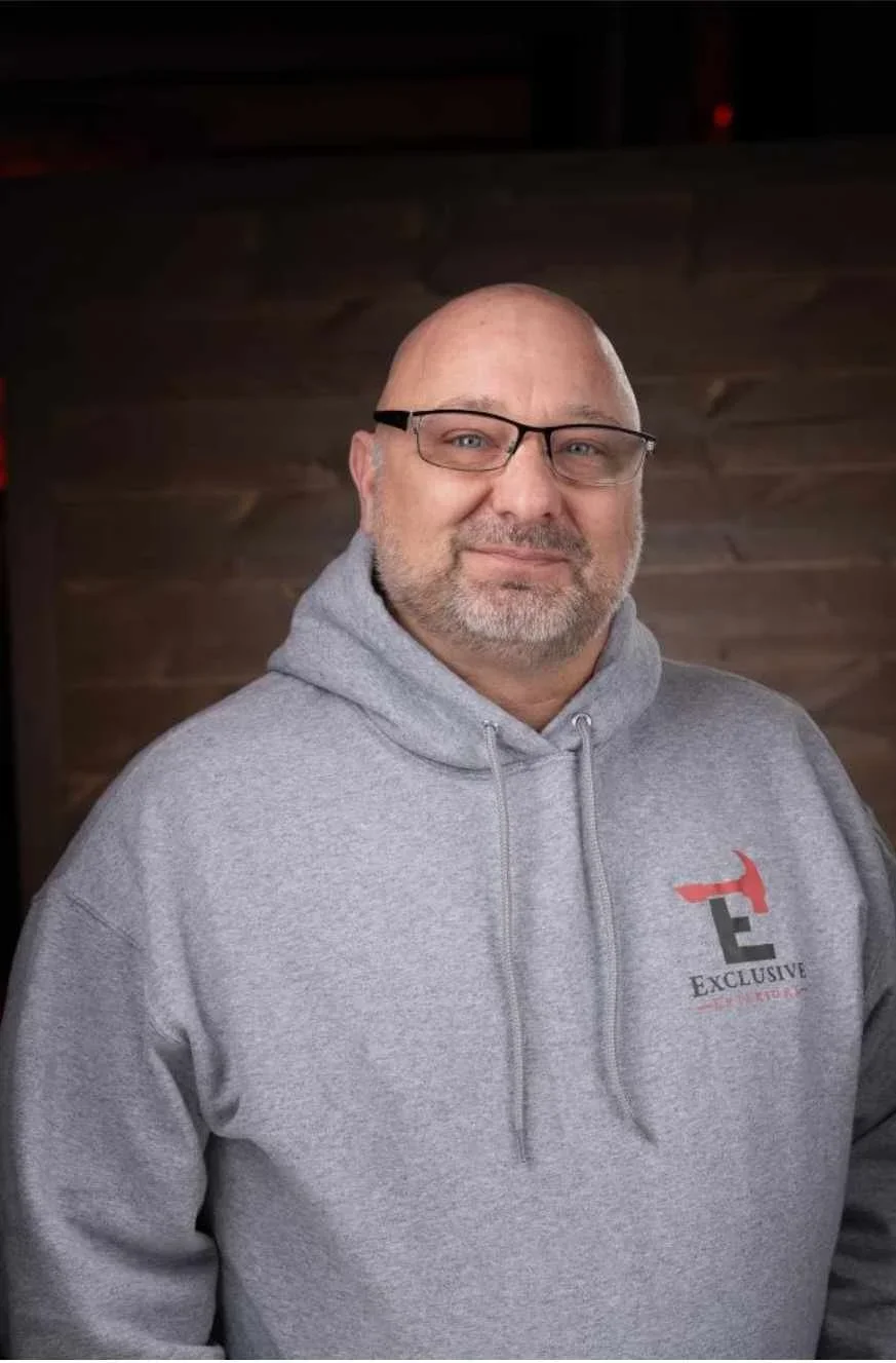A middle-aged man with glasses, a bald head, and a beard wearing a gray hoodie with the logo 'Exclusive' on the chest, standing in front of a wooden wall.