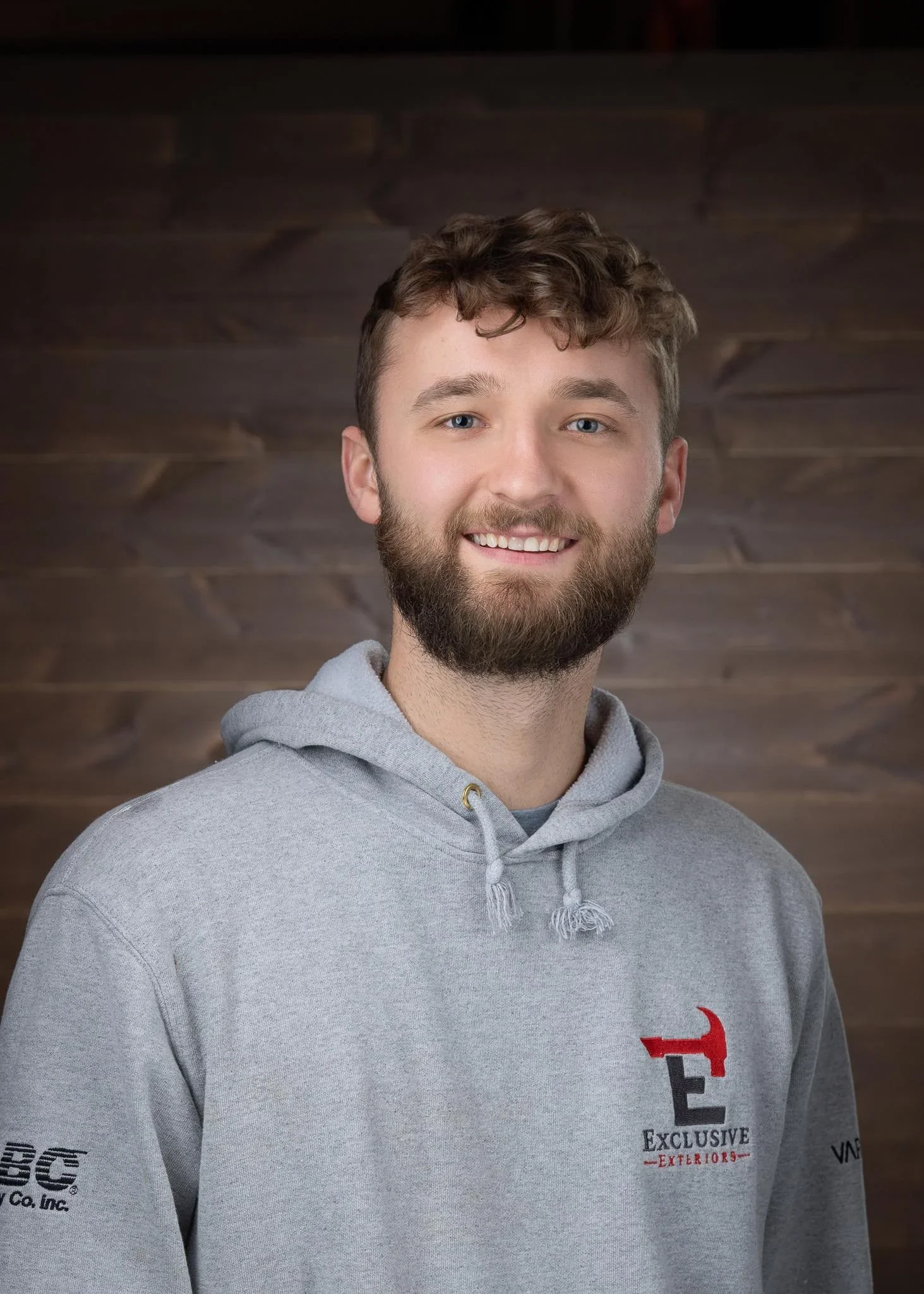 A young man with a beard and curly hair wearing a gray hoodie, smiling in front of a dark, wooden background.