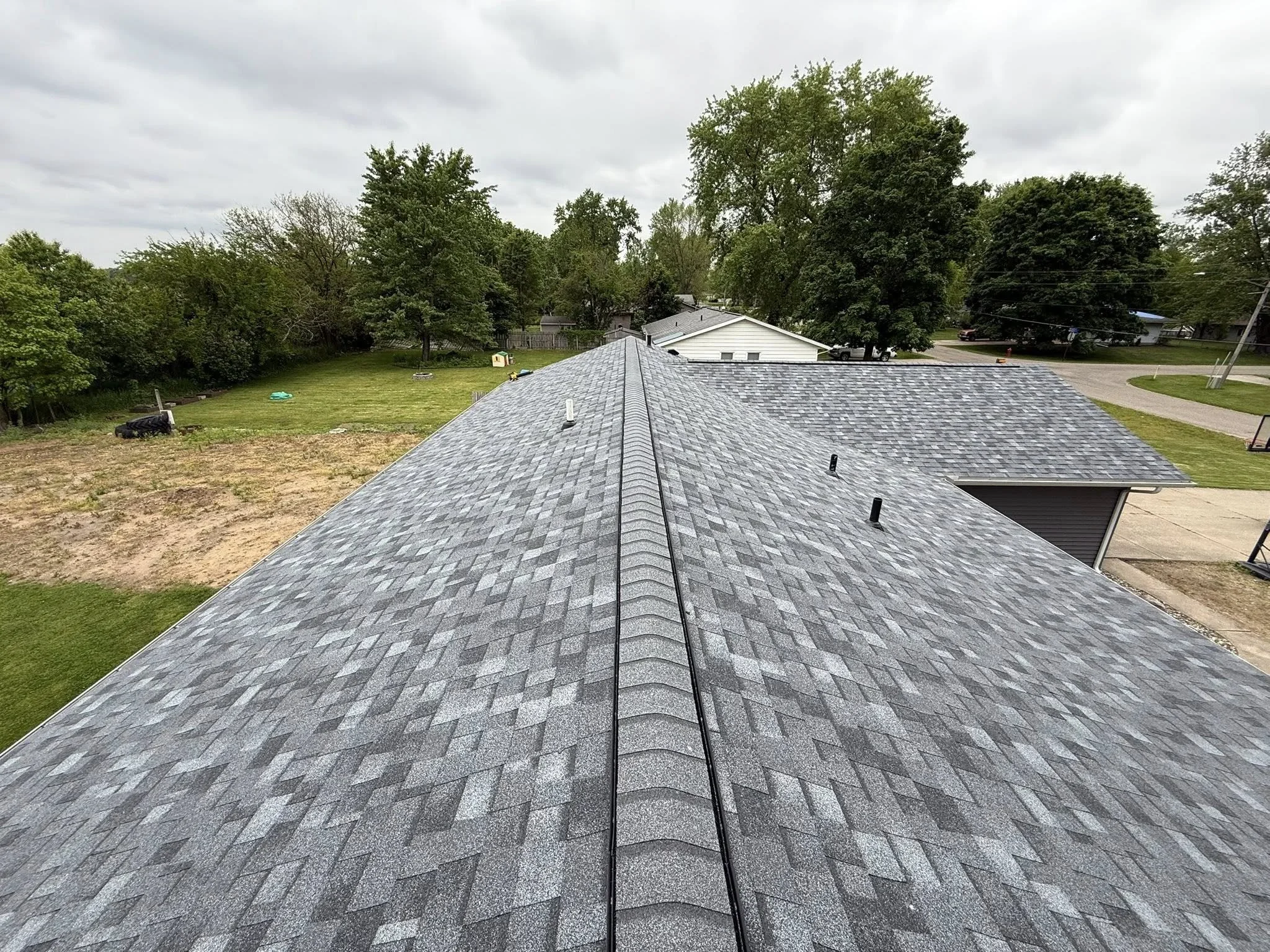 Aerial view of a house roof with gray shingles, surrounded by green trees and neighboring houses, under a cloudy sky.