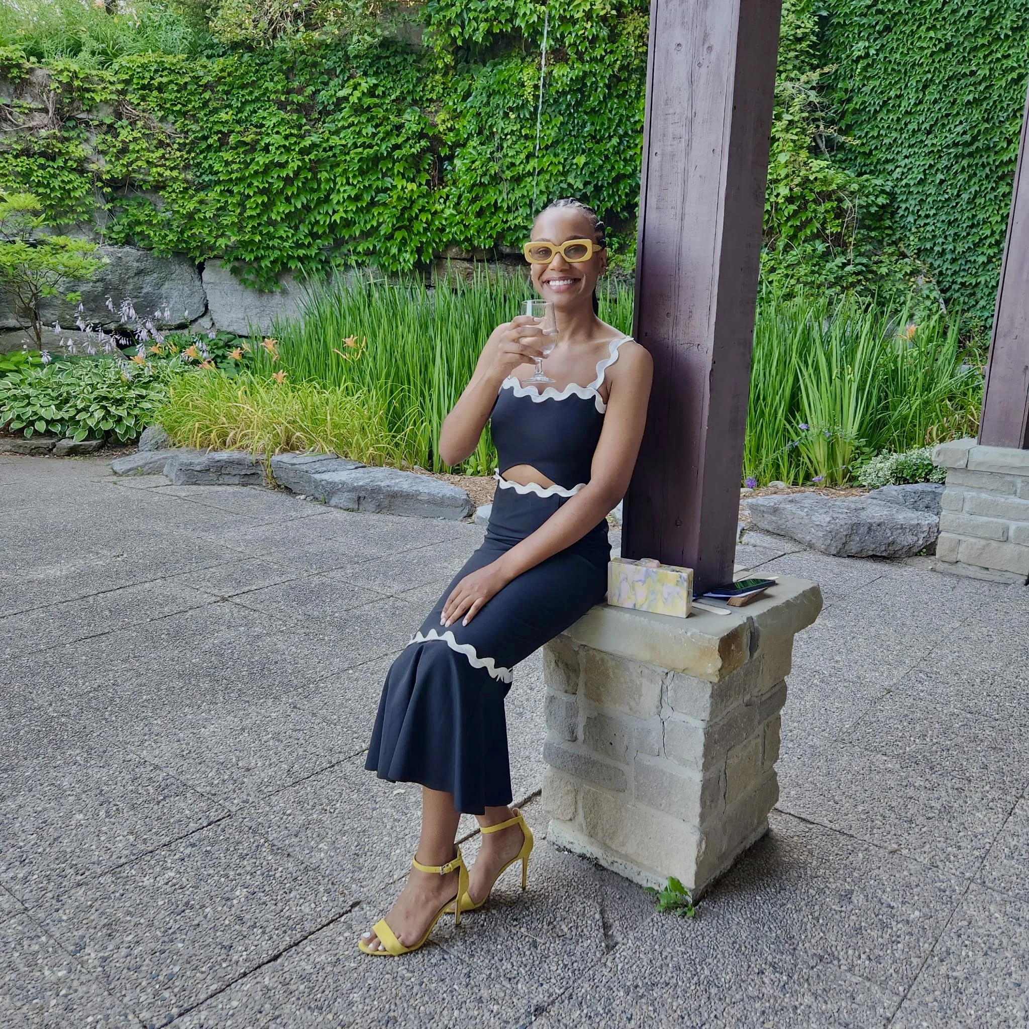 A woman wearing a black dress with white ruffle details, yellow high heels, and yellow glasses, sitting outside on a stone ledge, smiling and holding a glass of wine, with greenery and garden plants in the background.