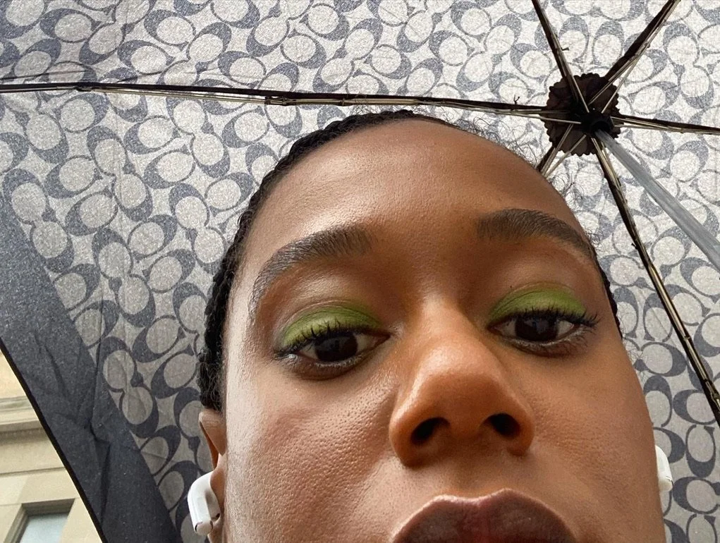 Close-up of a woman with green eyeshadow, holding an umbrella with a patterned interior, and wearing wireless earbuds.