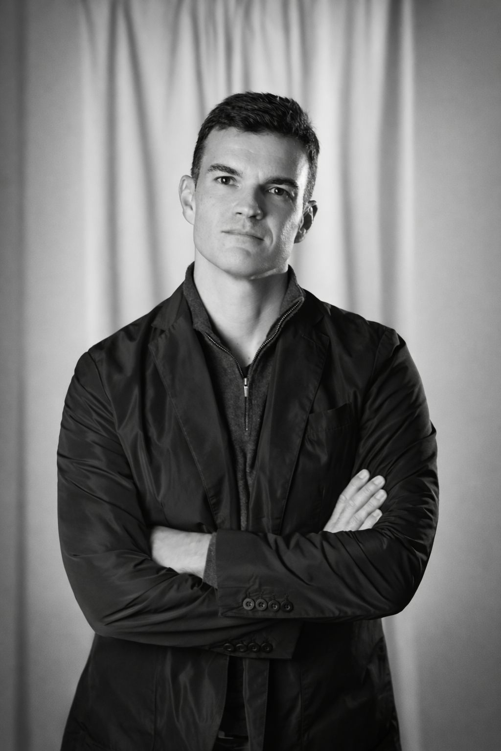 Retrato en blanco y negro de un hombre con expresión seria, de cabello oscuro y corto, con brazos cruzados, vistiendo chaqueta y fondo de cortinas.