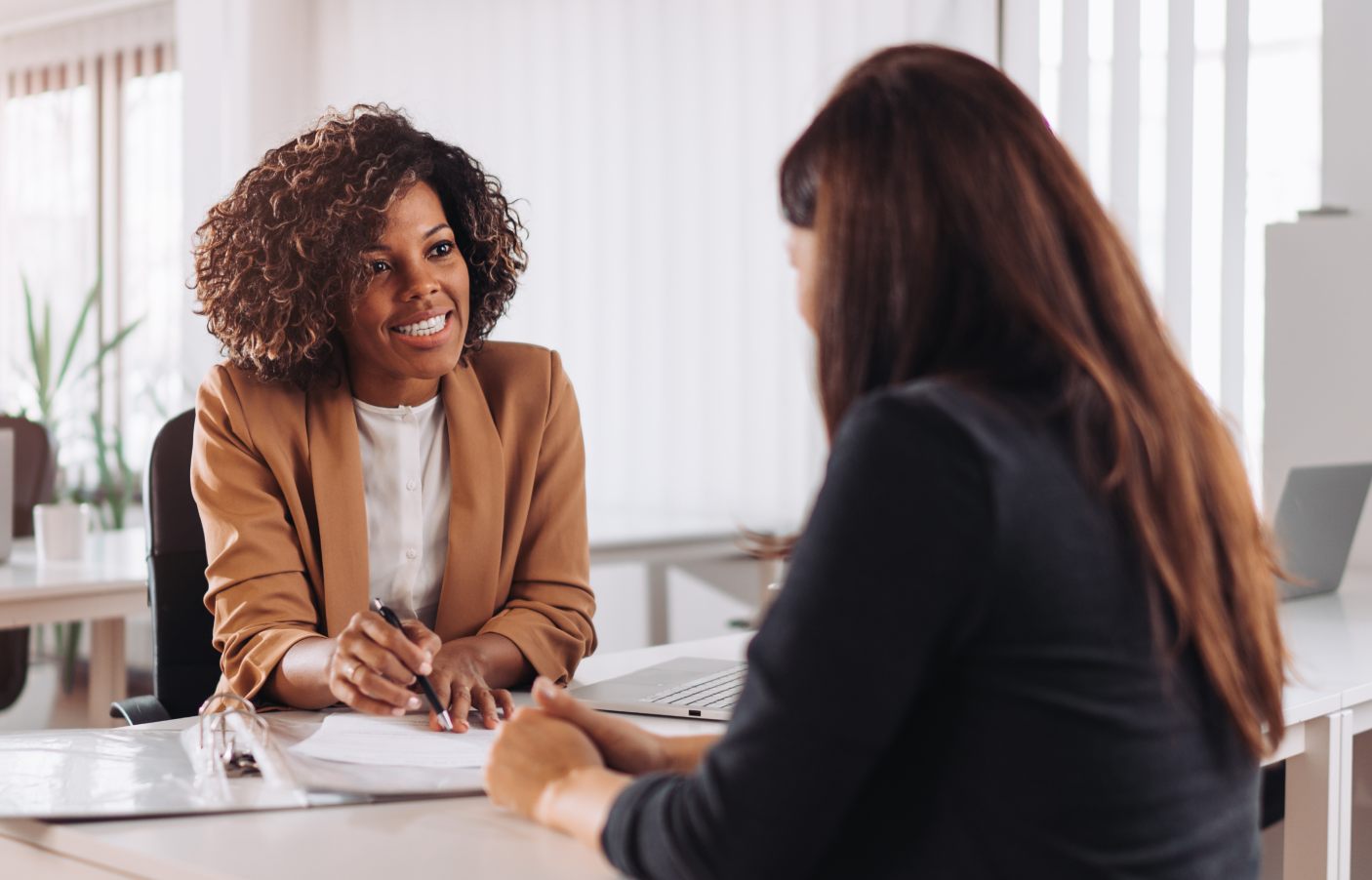 two women sitting facing each other at desk having a business conversation, and pointing at paperwork on desk