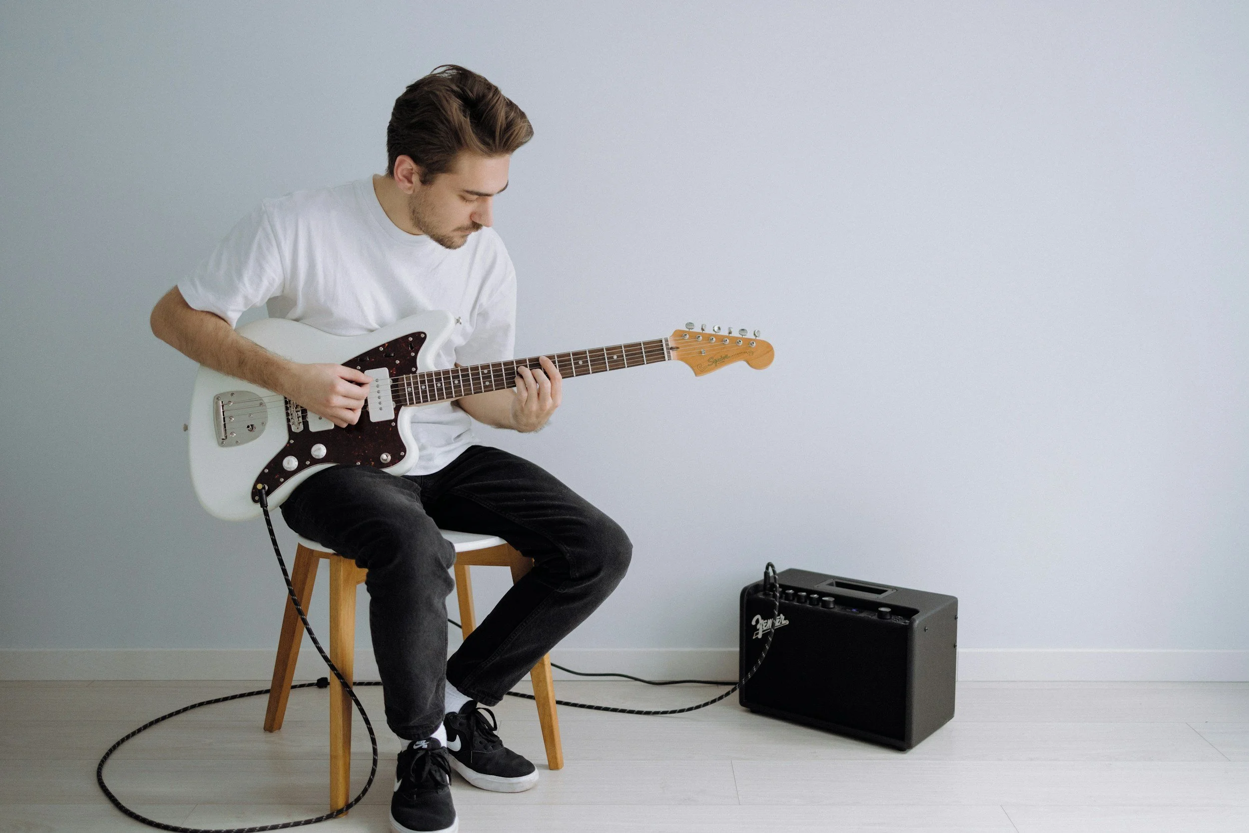 Un jeune homme assis sur une chaise jouant de la guitare électrique blanche, avec un amplificateur à côté sur le sol dans une pièce avec un mur blanc