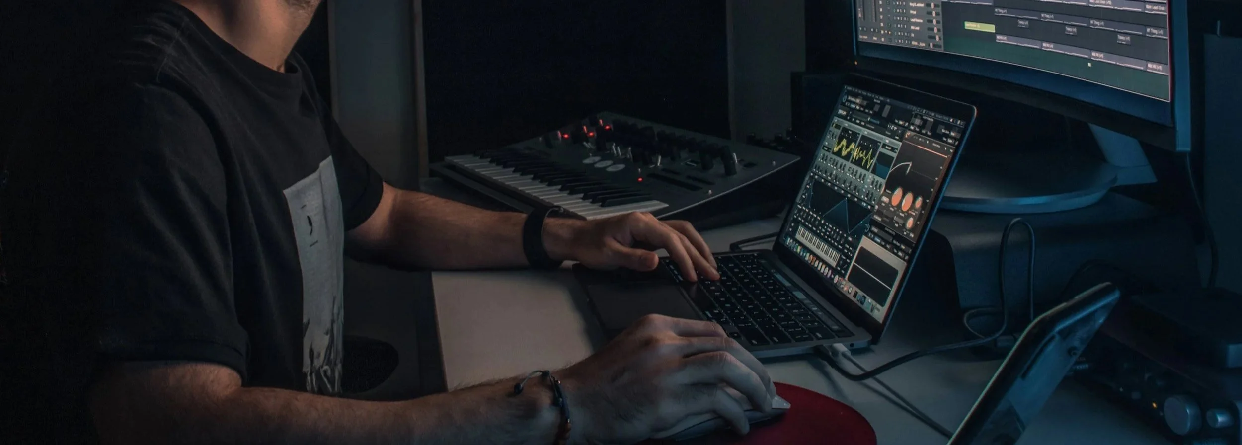 Personne utilisant un ordinateur portable avec un logiciel de production musicale, entouré d'équipements audio et d'écrans affichant des graphiques et des paramètres sonores.