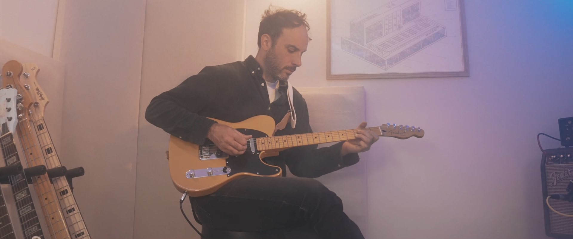 Homme jouant de la guitare électrique dans un studio avec plusieurs guitares en bois derrière lui et un ampli Fender à droite.