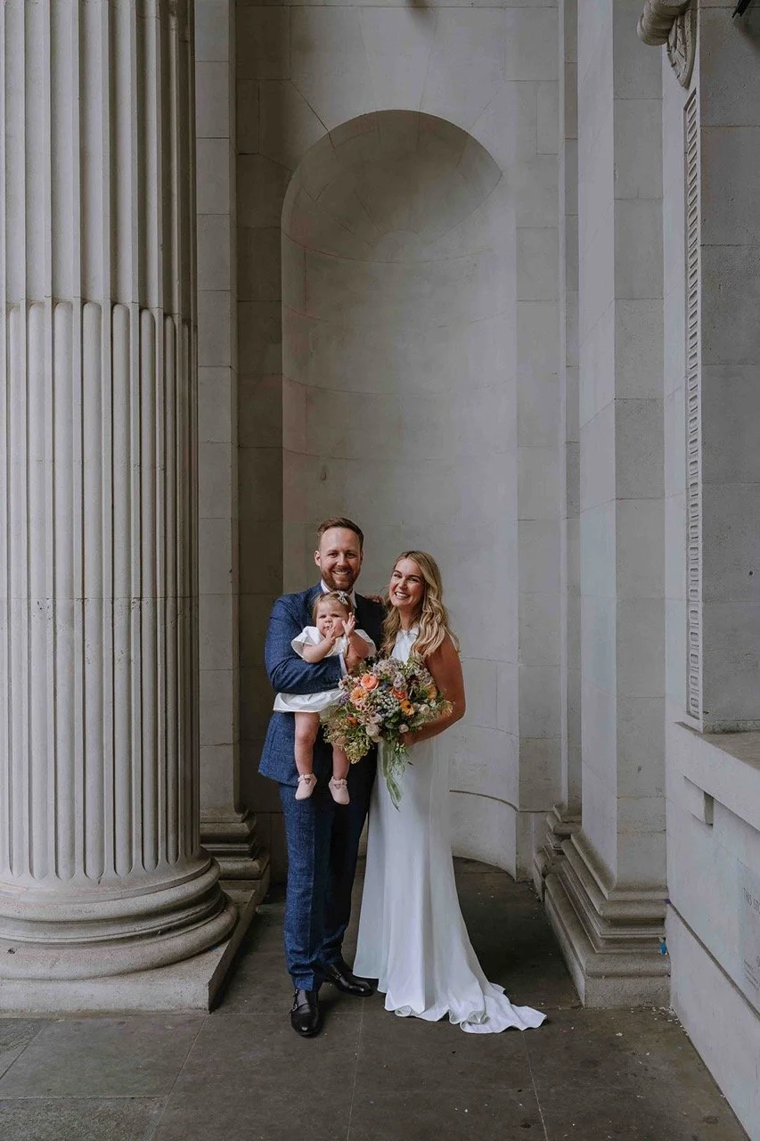 A happy family of three at a wedding, standing in front of classical columns. The man is in a blue suit, the woman in a white wedding dress holding a bouquet, and a young girl in a light dress, sitting on the man's arm.