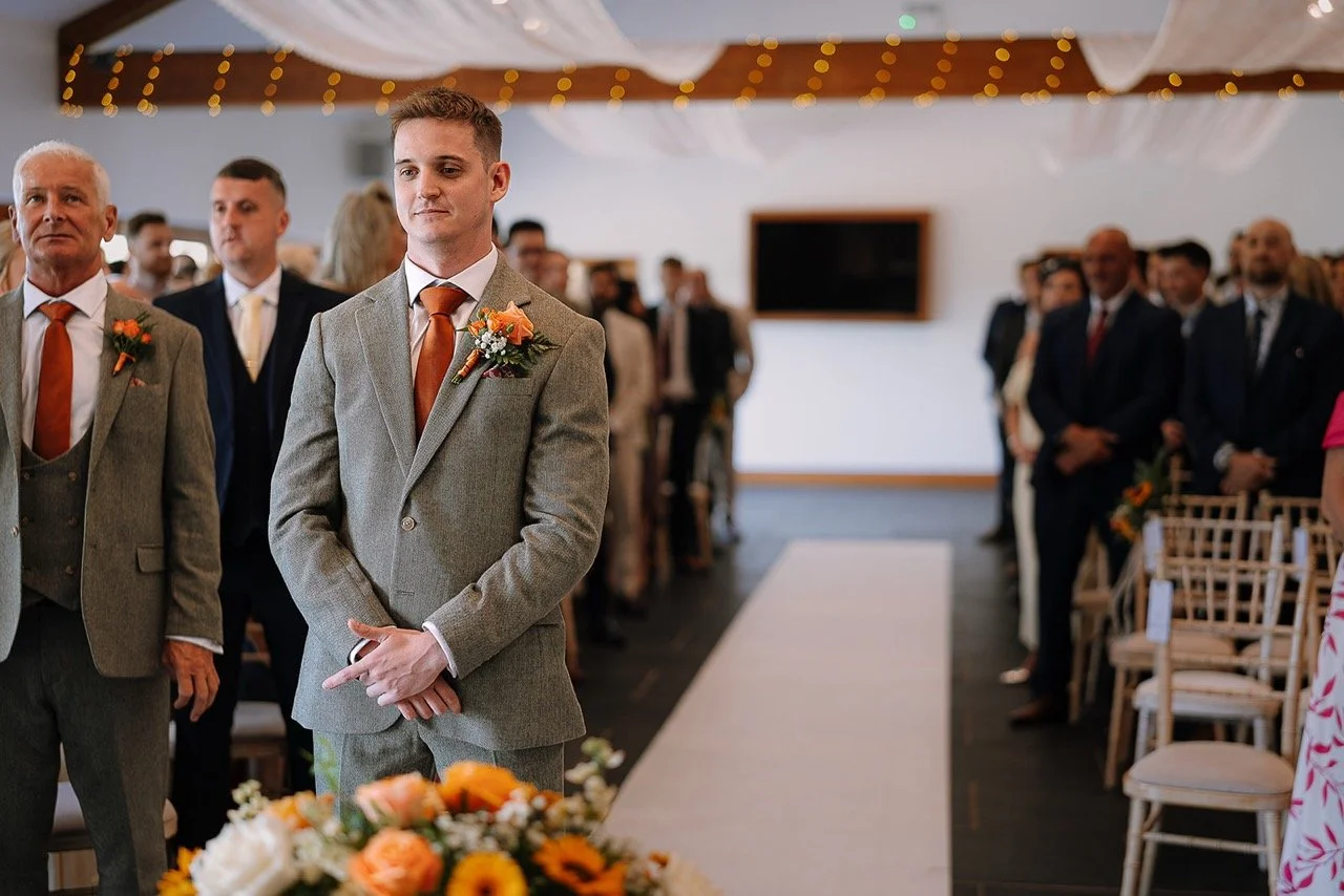 Groom standing at the altar during a wedding ceremony with guests seated behind him in a decorated hall.