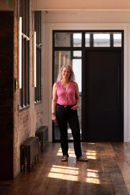 Woman standing inside a room with wood flooring, brick walls, large window, and black door, smiling at the camera.