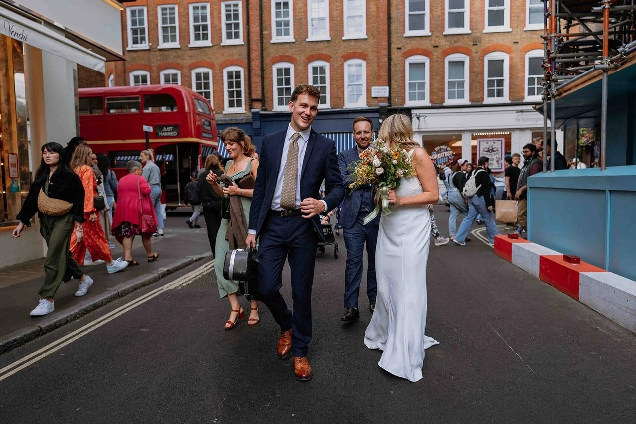 A newlywed couple walking on the street with a group of people and a red double-decker bus in the background.