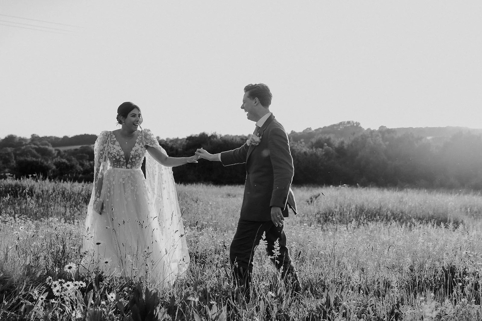 A bride and groom holding hands in a field of wildflowers, smiling at each other during sunset, in black and white.