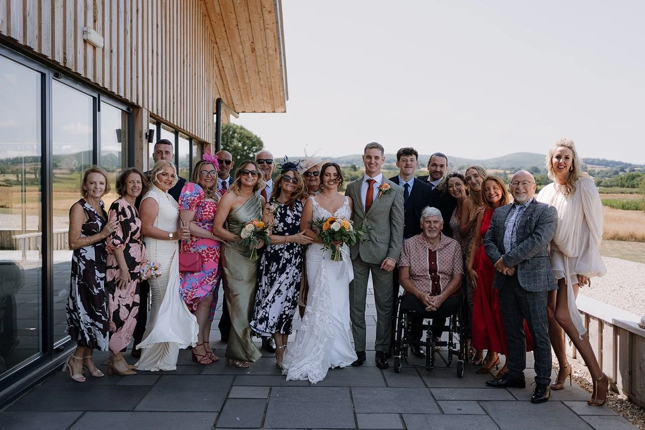 A large group of people, including a bride and groom, standing outdoors on a terrace during a wedding celebration. The bride is holding a bouquet, and some guests are also holding flowers. The group is smiling and dressed in formal and semi-formal at