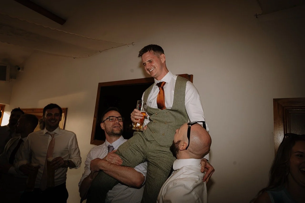 A man in a gray suit with a white shirt and orange tie is sitting on the shoulders of a man with glasses and a white shirt, holding a beer, smiling and enjoying himself at a social event. Several other people are in the background, some smiling and w