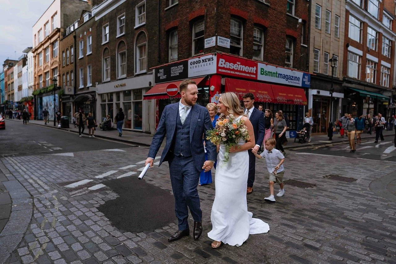 A bride and groom walking hand in hand across a city street during their wedding celebration, surrounded by pedestrians and city buildings.