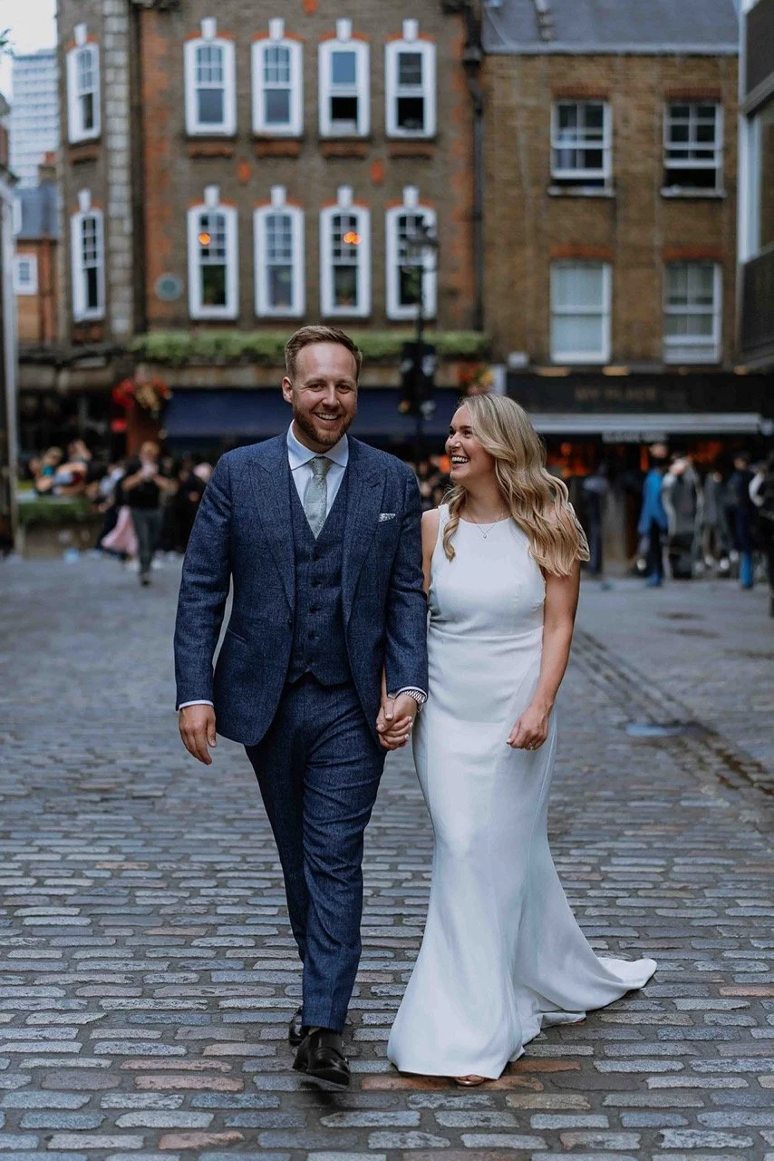A happy couple, dressed in wedding attire, walking hand-in-hand on a cobblestone street in an urban setting, smiling and looking at each other.