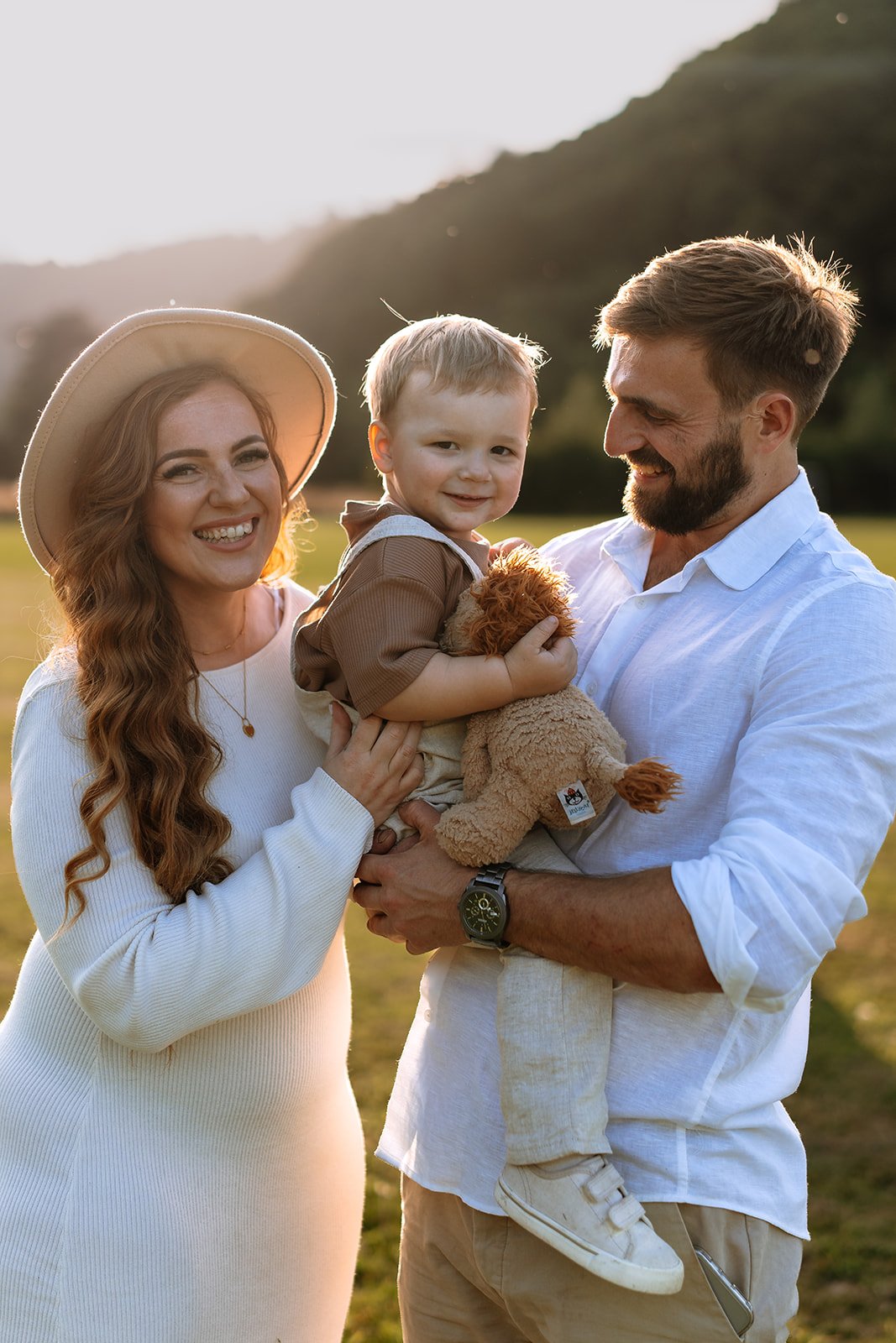 A smiling family of three, including a woman with long red hair wearing a hat, a man with a beard, and a young boy holding a stuffed animal, standing outdoors during sunset with mountains in the background.