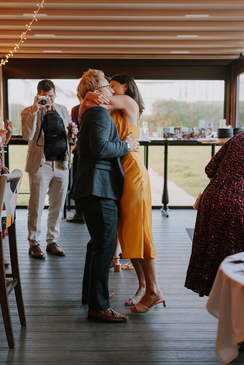 Two women, one in a floral dress and the other in a yellow dress, share a hug at a social gathering. Background shows other guests, a photographer, and a window with a view outside.