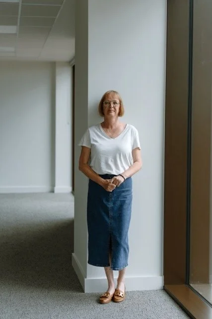 A woman with short red hair and glasses standing indoors near a window, wearing a white t-shirt, blue skirt, and sandals.