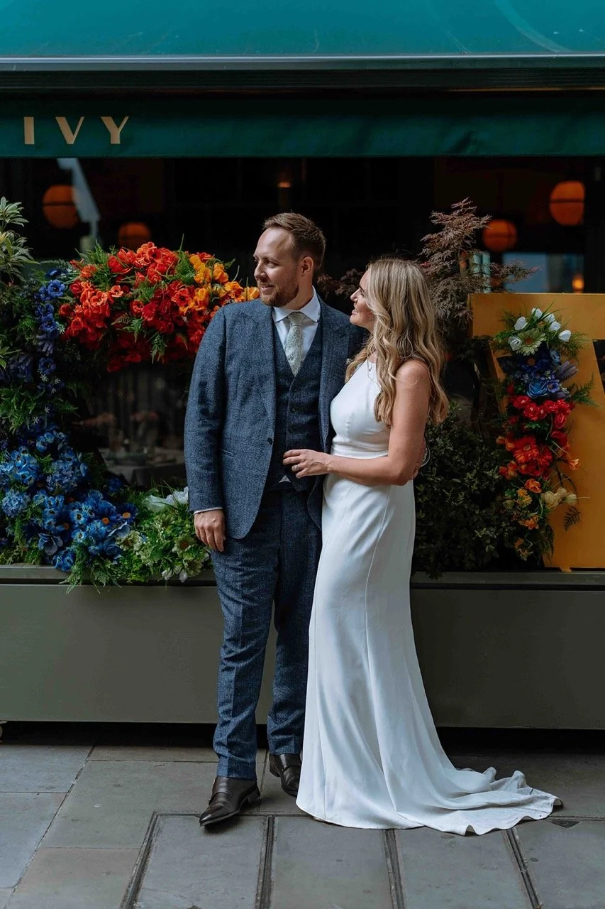 A couple dressed in formal wedding attire standing in front of a floral display with red and blue flowers. The man is wearing a gray suit with a vest and tie, and the woman is wearing a white sleeveless wedding gown. They are standing outdoors, near 