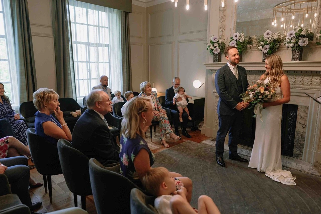 A wedding ceremony with a bride and groom standing in front of the fireplace, surrounded by seated guests. The bride holds a bouquet and is wearing a white dress, while the groom wears a dark suit. The room is elegant with large windows, floral arran