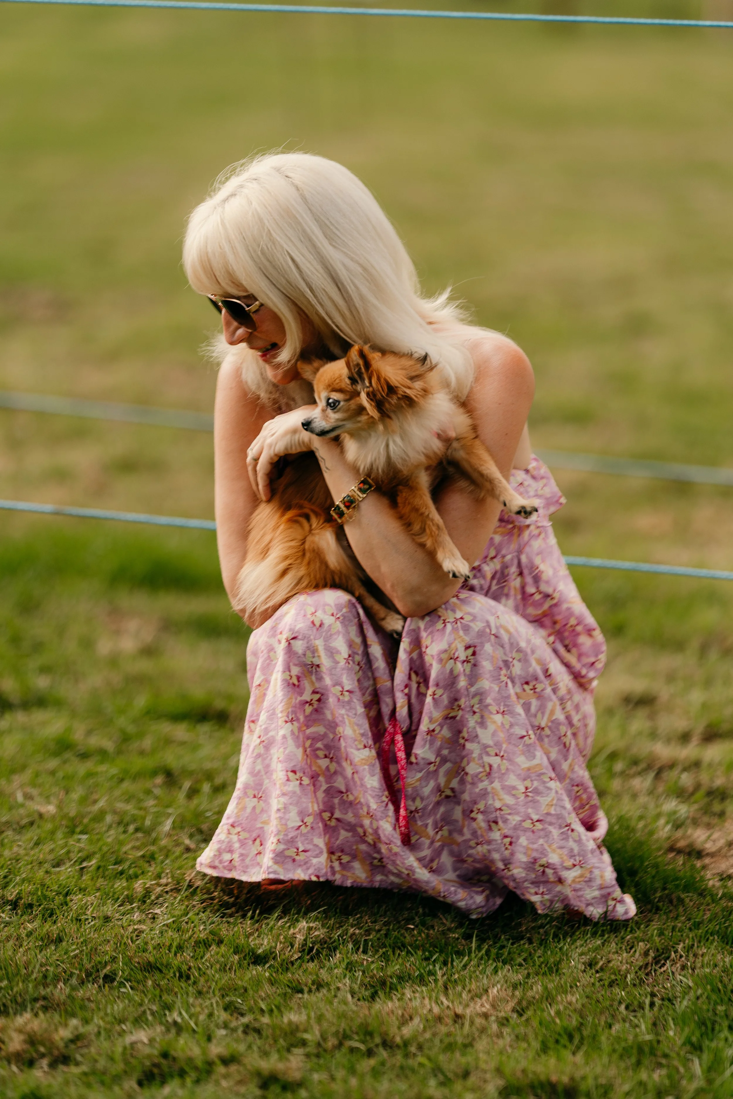 Woman with blonde hair wearing glasses and a pink floral dress crouching on grass, holding and cuddling a small brown dog outdoors.