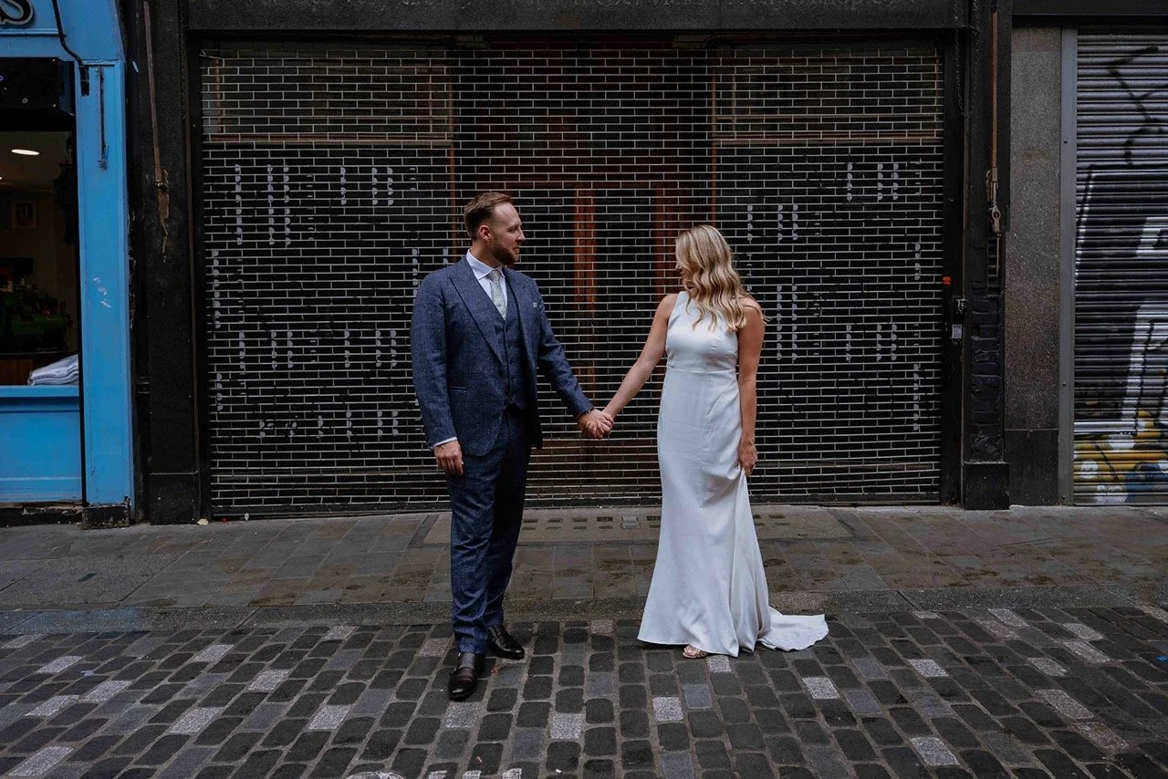 A man in a blue suit holding hands with a woman in a white wedding dress standing on a cobblestone street in front of a metal gate.