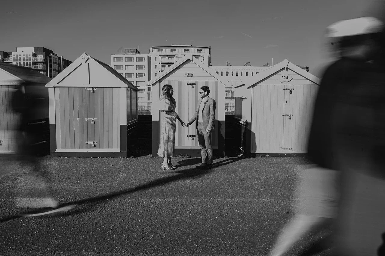 A black and white photo of a couple holding hands underneath a small house with accompanying persons blurred in motion on the sides.
