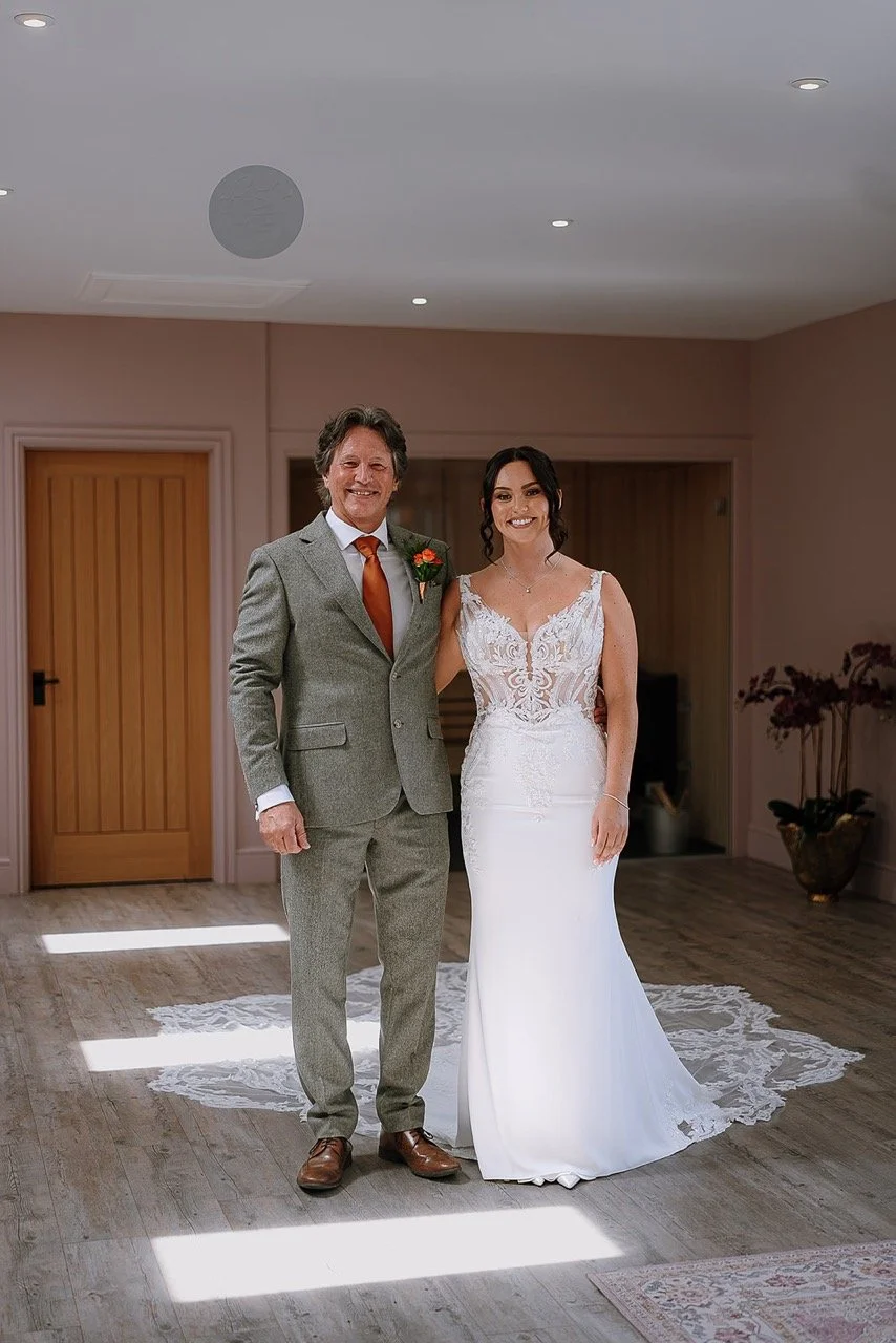 Man and woman in wedding attire standing indoors, smiling, with a wooden door and plant in the background.