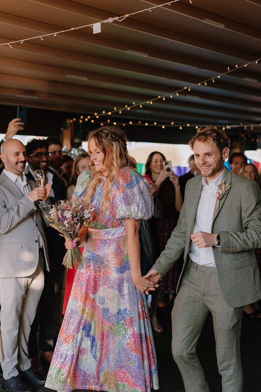 A happy bride and groom holding hands during their wedding reception with guests smiling and taking photos in the background under a decorated ceiling.