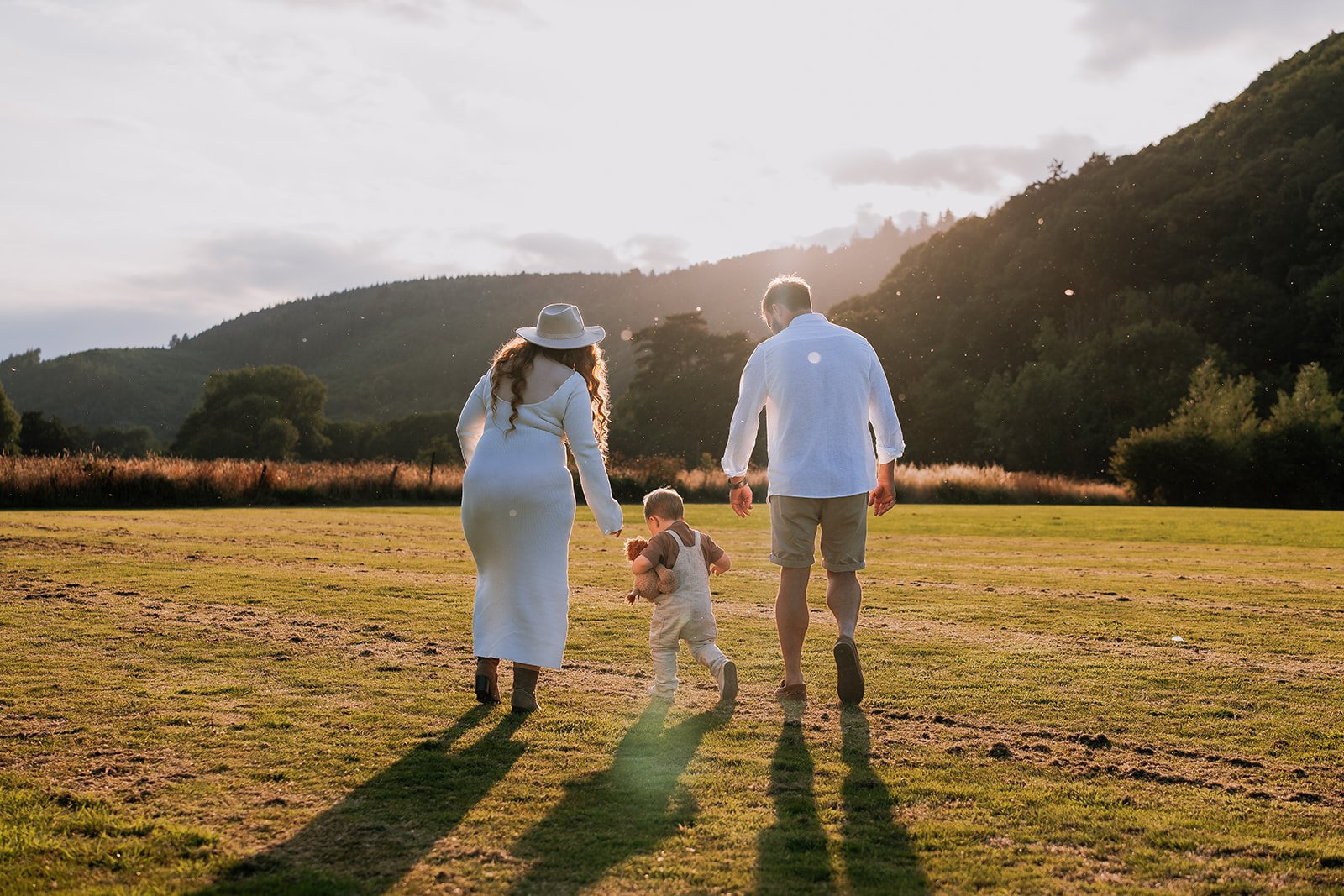 A family of three walking across a grassy field during sunset, with mountains and trees in the background.